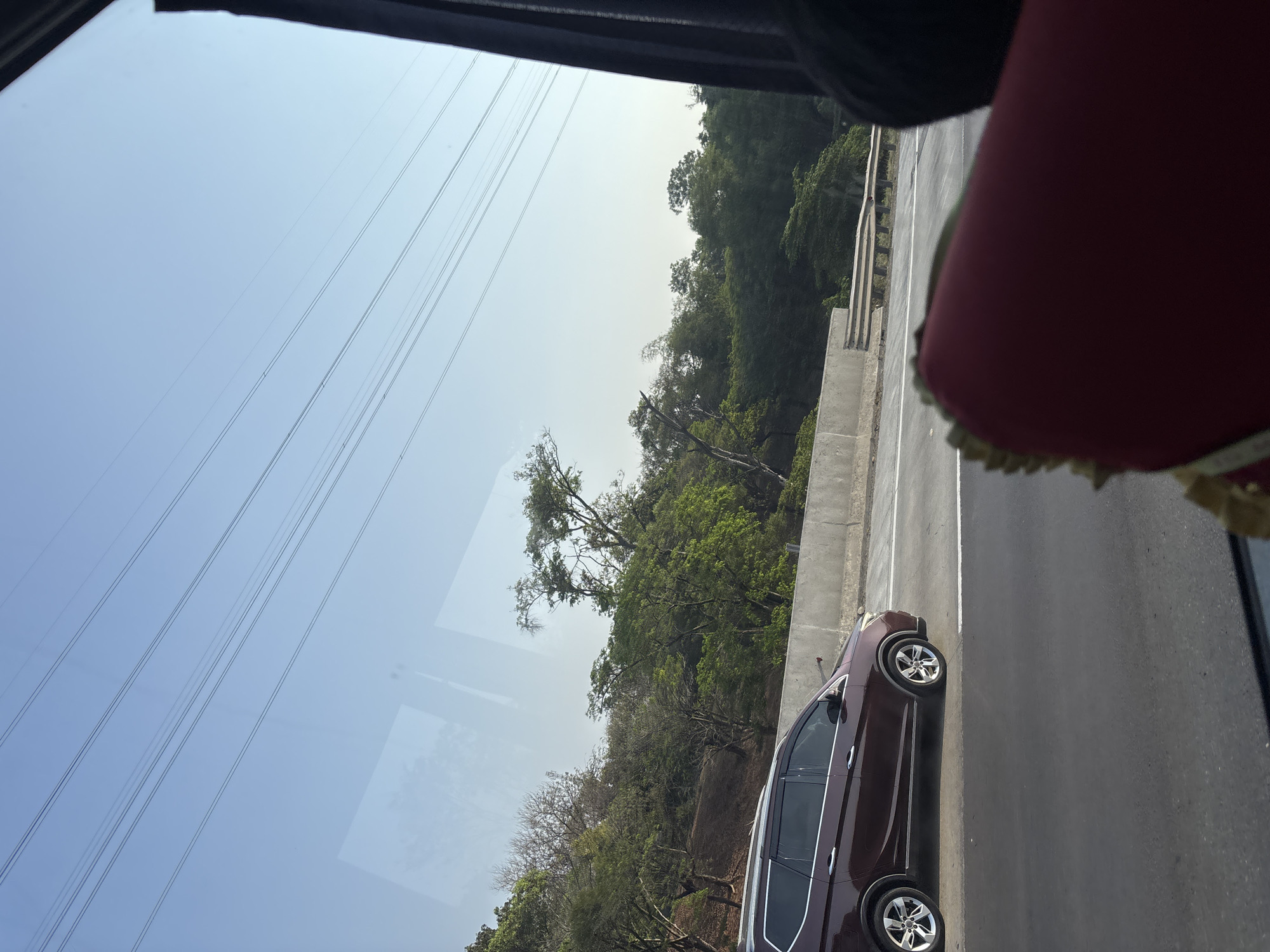 The image, captured from inside a vehicle on a highway in Karawang, Indonesia, shows a maroon SUV driving on the asphalt road. The SUV is positioned on the left side of the frame, in the middle lane. The road is bordered by a concrete barrier and a lush green hillside covered with trees. In the sky, several parallel power lines stretch across the frame, indicative of the highway infrastructure. The foreground is partially obscured by a red upholstered object, possibly part of the vehicle's interior. The lighting suggests it is daytime, with a clear sky.