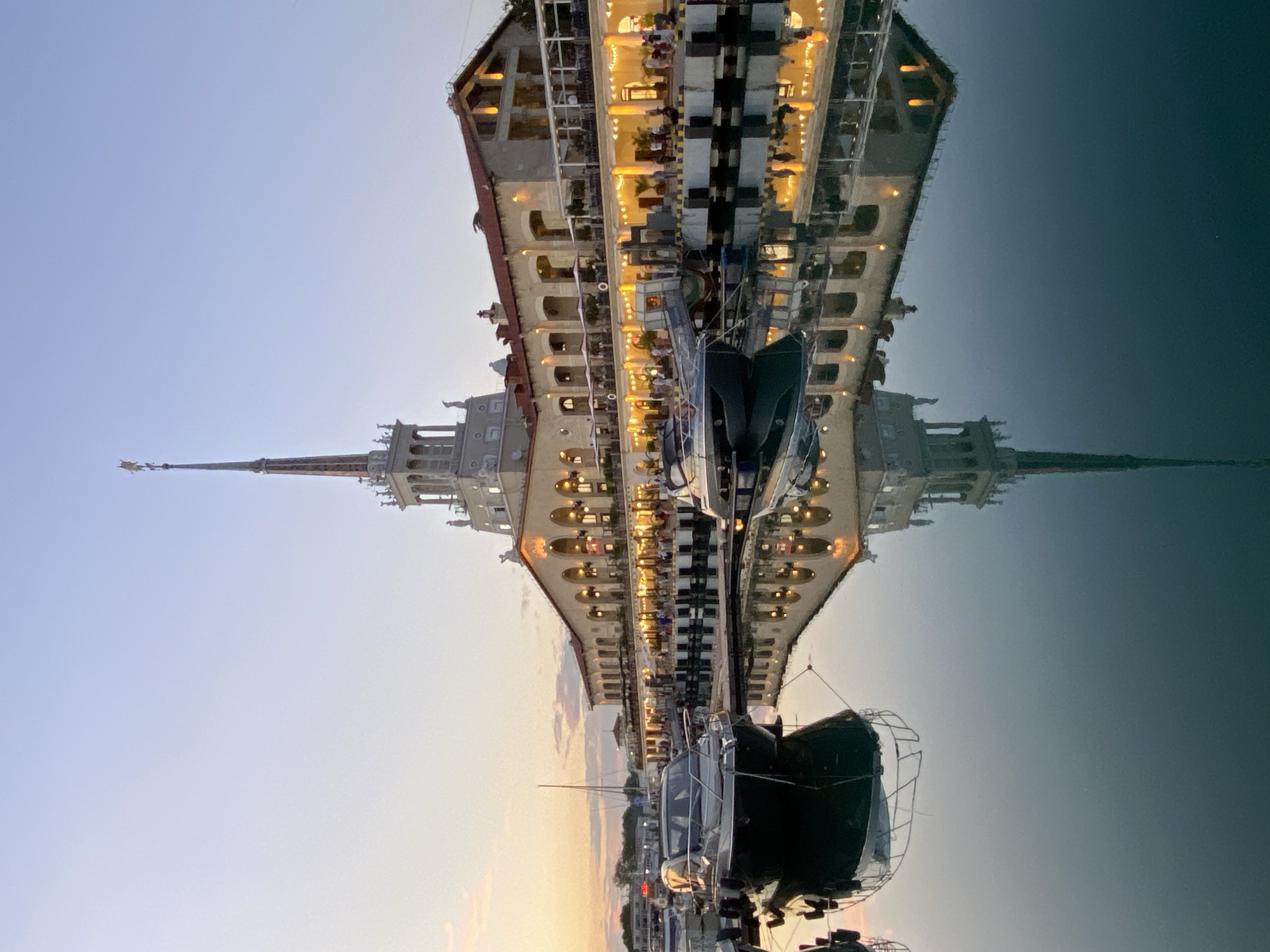 This is a vertical, eye-level, long shot of the Sochi Sea Port building, with boats in the foreground on a calm body of water reflecting the scene. The building is large, pale yellow, with red-tiled roofs and a prominent central tower topped with a golden star. The building has a colonnaded walkway with many arched openings, lit by warm lights. Small tables with umbrellas are visible outside the walkway. Several boats are docked in the water, the closest one being a dark gray motorboat. The sky is a pale blue fading to a soft orange near the horizon, suggesting either dawn or dusk. The water is dark and reflects the building, the boats, and the sky with clarity. There are a few people visible on the walkway and dock, appearing as small silhouettes. The architectural style of the building is grand and somewhat imposing.