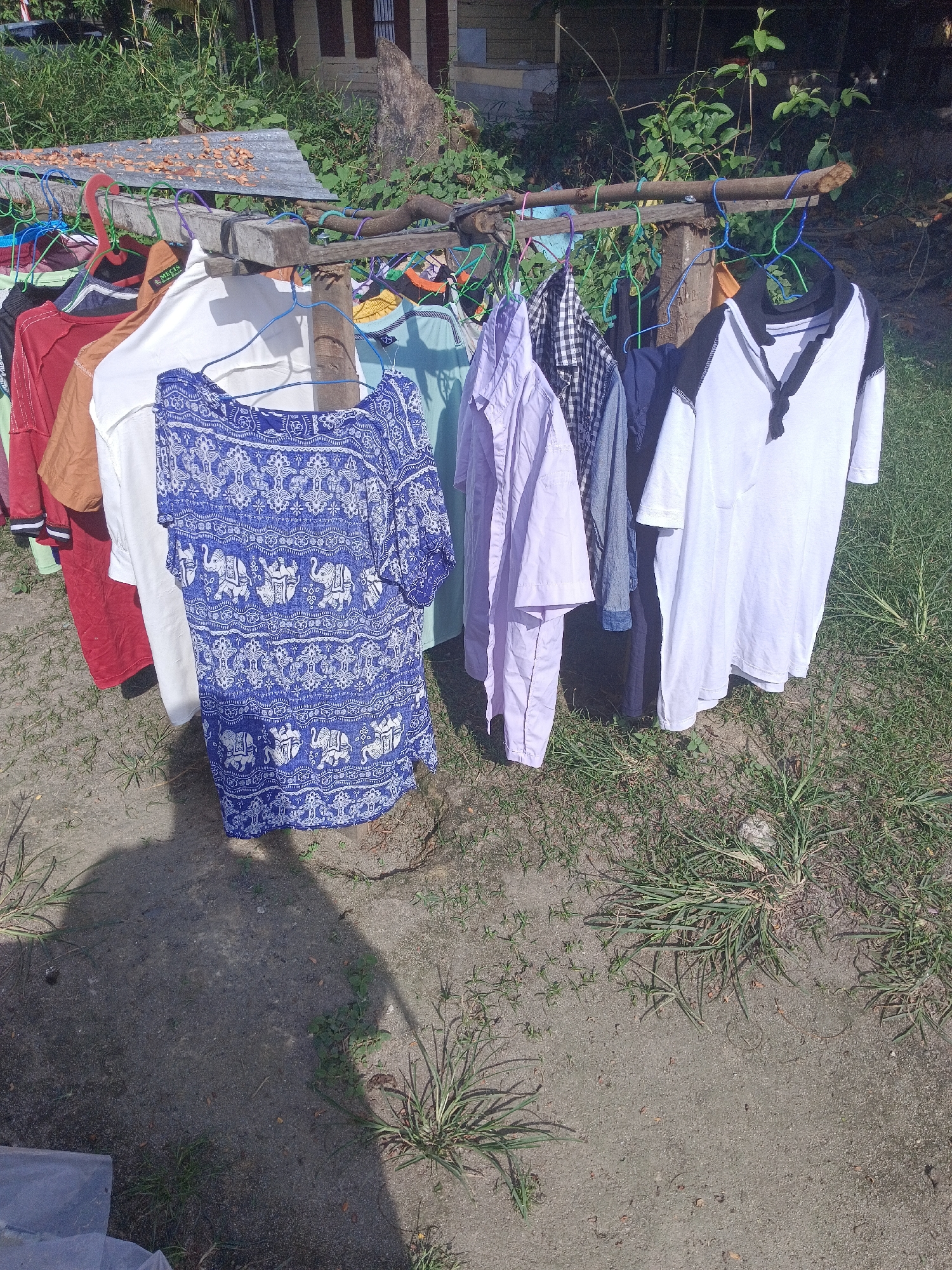 In the city of Aek Kanopan, Indonesia, an outdoor clothesline displays a variety of garments. The clothes are hung on colorful plastic hangers, strung across a makeshift rack constructed from branches. Among the items are a blue and white patterned shirt, a white t-shirt with black trim on the collar and sleeves, a checkered shirt, a light purple long-sleeved shirt, and several other t-shirts in red, brown, and white. The background features lush green foliage, a portion of a building with dark-colored window frames, and a weathered, upright piece of wood. The ground beneath the clothesline is a mix of dry soil and sparse grass. Sunlight casts shadows on the ground, indicating a bright, daytime scene.
