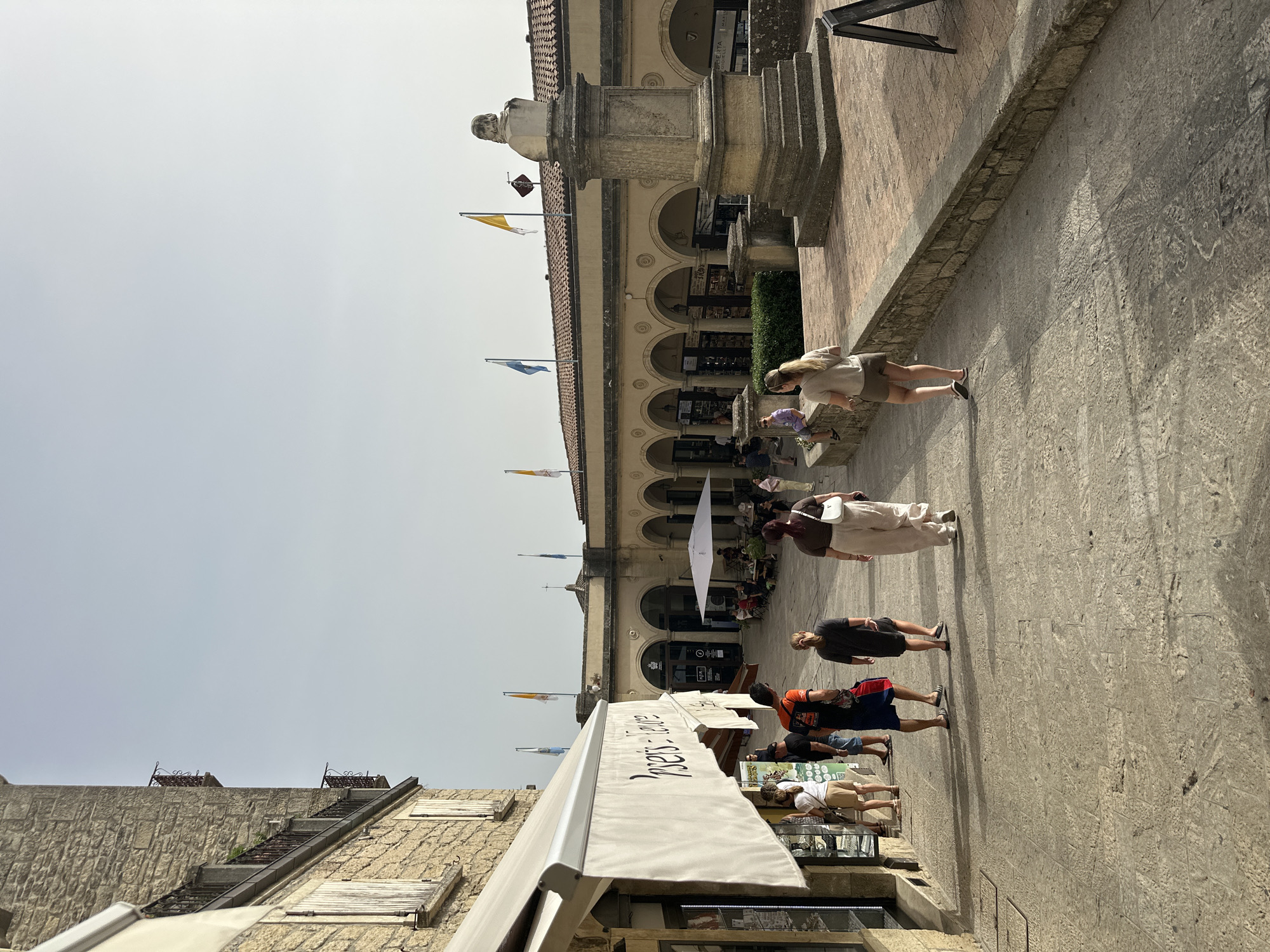 The image depicts a street scene in the City of San Marino, San Marino. In the foreground, a paved pathway runs diagonally from the lower left to the upper right. Several individuals are walking along this pathway. On the left side of the pathway, a building with a stone facade features multiple windows with shutters and a white awning extended over a storefront. A sign on the awning reads "orsetti - faure".
Further into the scene, the pathway opens into a wider area with a paved courtyard. To the right of this courtyard stands a stone monument topped with a bust. Behind the courtyard is a long building with a series of arched walkways. Various flags, including blue, yellow, and white, are visible flying above the building. In the courtyard, tables and chairs are set up, with some people seated. The sky above is clear and pale. The architecture suggests a historic European setting.