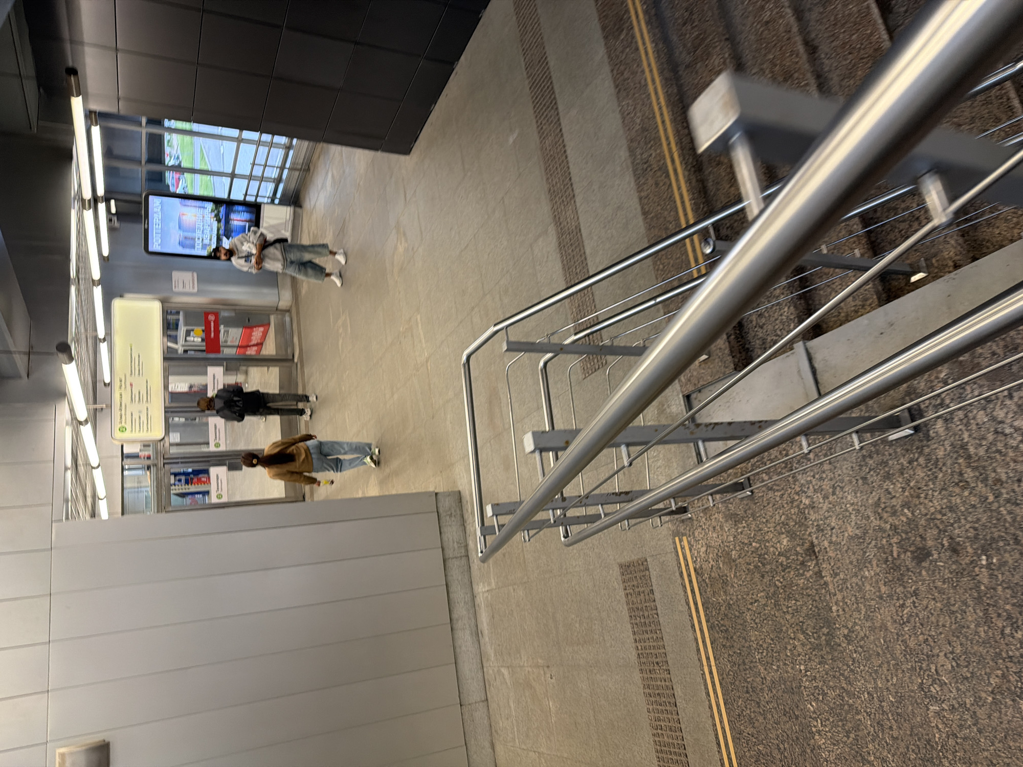 The image shows an interior view of a modern, well-lit public space, possibly a subway station or transit hub. In the foreground, a set of metal handrails and stairs are visible, suggesting a descent into the lower level. The floor is paved with large, textured tiles.
Further back, three individuals are present. On the left, a woman wearing a brown jacket and light blue jeans is walking away from the camera, holding a yellow object in her right hand. In the middle, a person in a dark jacket and dark pants is walking towards a set of glass doors. To the right, a man with a dark cap, grey hoodie, and light blue wide-leg pants stands looking at his phone. He is positioned in front of a large digital display showing an image of buildings labeled "ROTTERDAM" and text in Cyrillic script.
The environment features clean lines, metal accents, and bright fluorescent lighting. Behind the people, more glass doors lead to an outdoor area with greenery and parked cars visible through the windows. The overall impression is one of a contemporary, functional public transit facility.