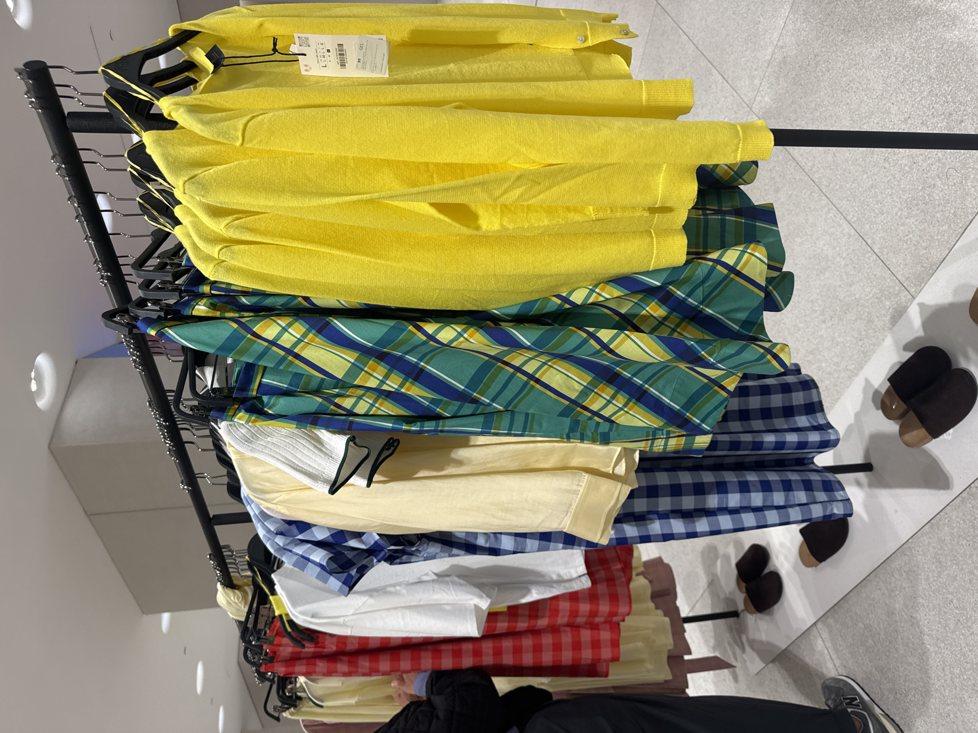 The image shows a clothing rack in what appears to be a retail store. The rack is filled with folded clothes, including yellow sweaters, plaid shirts in blue, green, and yellow, light yellow shirts, blue-and-white checkered shirts, white shirts, and red checkered shirts. The floor is a light grey tile. Two pairs of brown and black slippers are seen on the floor, and part of a person wearing black pants and a grey New Balance shoe is visible at the bottom of the frame. It is not possible to pinpoint the precise location in Batumi, Georgia from the information provided.