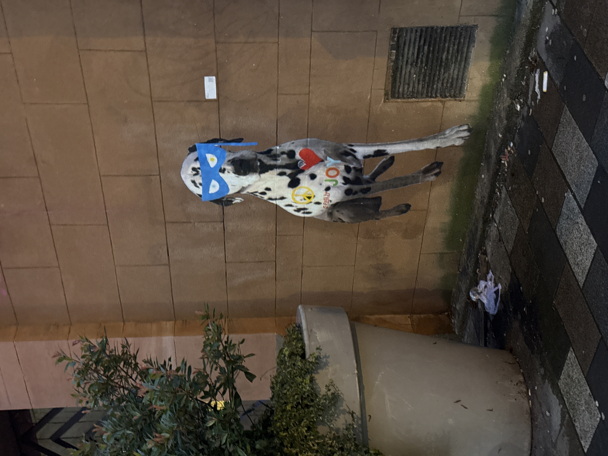 The image depicts a life-size cardboard cutout of a Dalmatian dog wearing a blue superhero mask, adhered to a brick wall in Glasgow, United Kingdom. The dog's cutout is adorned with a peace symbol on its left side and a red heart and the word "JOY" on its right. The wall is made of large, tan-colored rectangular bricks. To the left of the cutout, a large, grey planter contains dense green foliage. The ground in front of the wall is paved with interlocking square tiles and appears wet, with some litter scattered on it, including cigarette butts and a crumpled piece of paper. A metal grate is visible on the wall to the right of the dog cutout. The lighting suggests it is nighttime or dusk, with artificial light illuminating the scene.