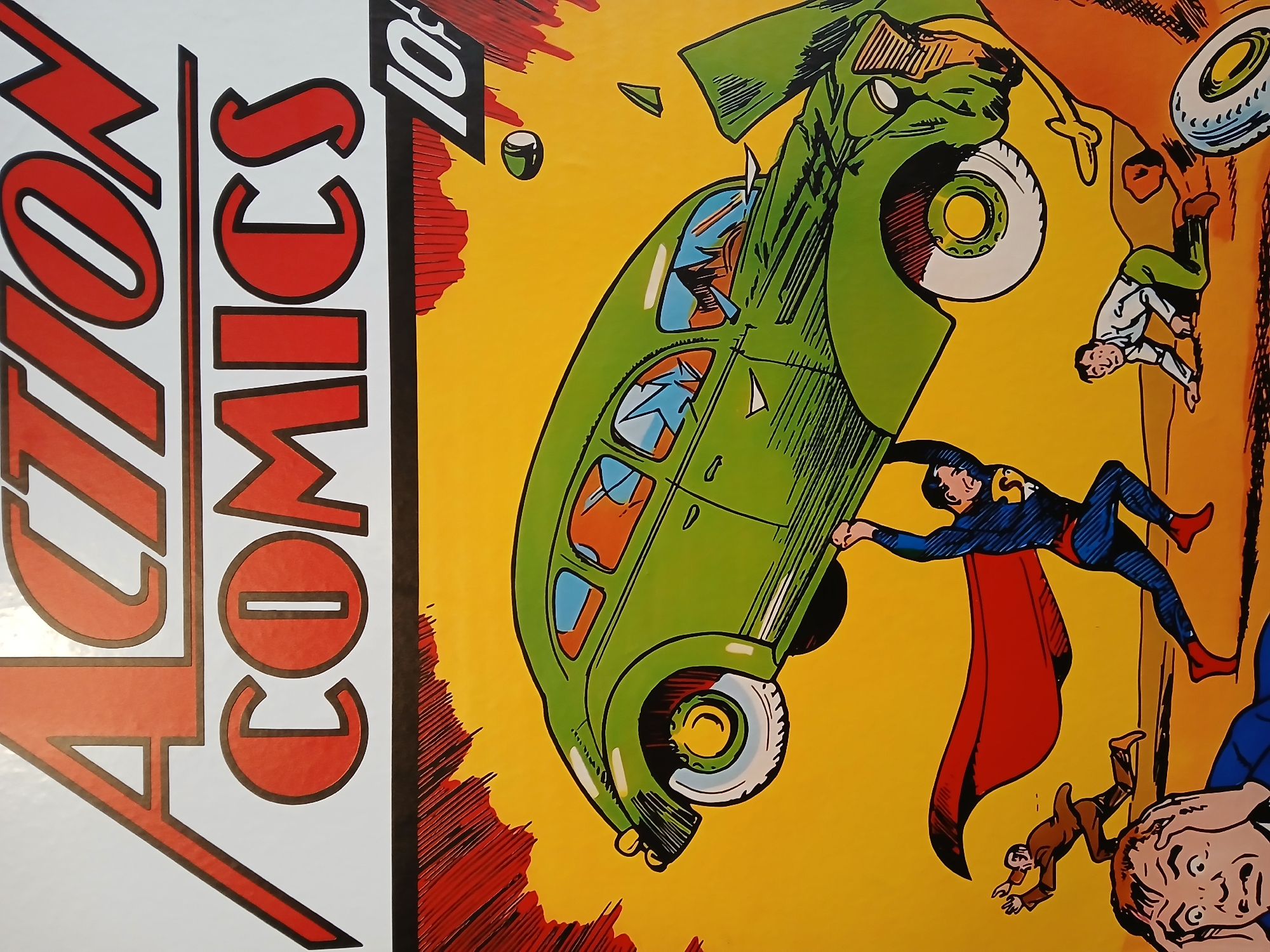 The image shows a reproduction of the cover of Action Comics #1, featuring Superman lifting a green car above his head. People are being thrown from the vehicle. The background is yellow with red and black stylistic markings. The "Action Comics" title is prominently displayed in red lettering on a white background with black outlining on the left side of the image. A price of "10¢" is displayed at the top-right. The location of Porlamar, Venezuela is not discernible from the image.