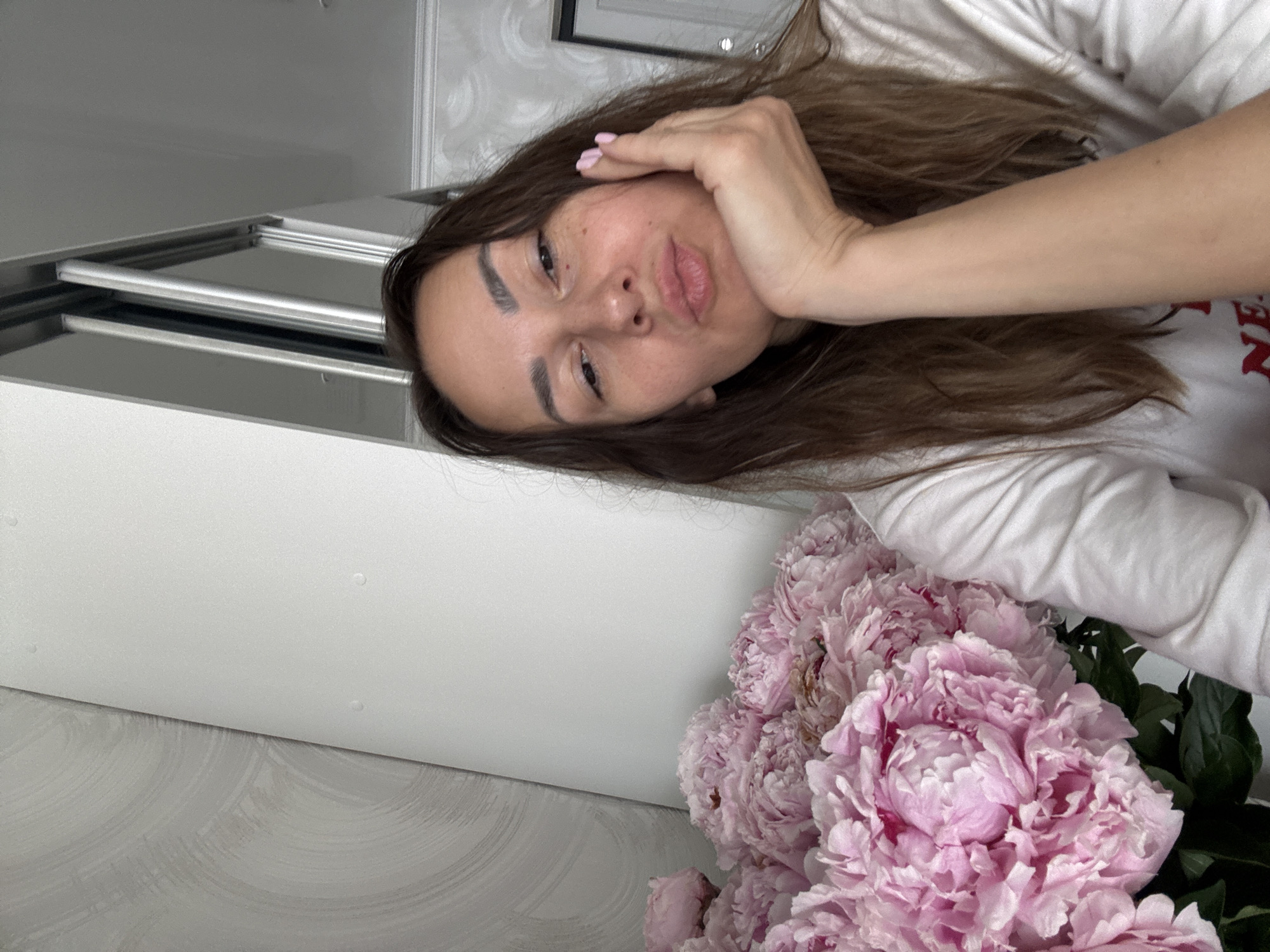 The image shows a selfie of a woman in Sochi, Russia. She is indoors, with a bouquet of pink peonies in the foreground. The woman is leaning her head on her hand, and she has a pouty expression. The background is a plain white wall with a subtle textured pattern. The overall impression is one of casual intimacy and aesthetic appreciation.