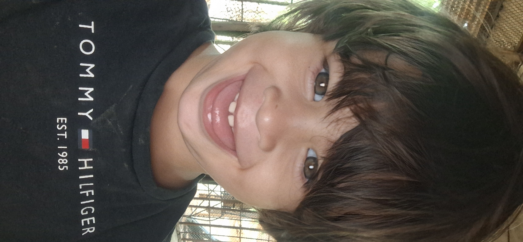 A young child, likely a toddler, is the central subject of this close-up shot. The child is grinning broadly, mouth open to reveal teeth. The child has brown eyes and dark brown hair that falls across the forehead. The child is wearing a dark-colored t-shirt featuring the "TOMMY HILFIGER EST. 1985" logo. The photograph appears to have been taken in an outdoor setting. The exact location in the city of Vallecito, Venezuela is impossible to know from the image provided.