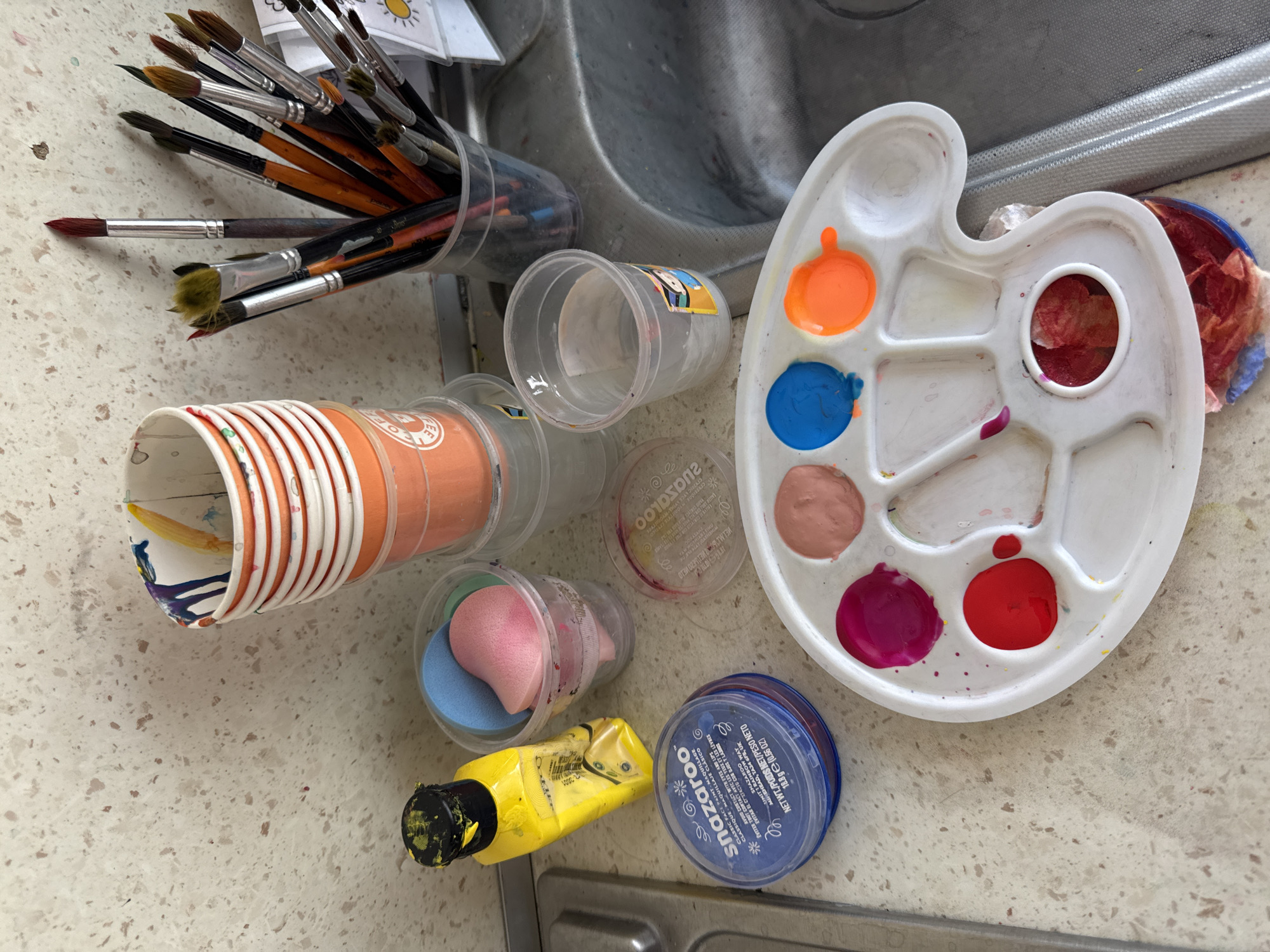 A still life composition depicts art supplies arranged on a countertop. In the upper right, a cluster of paintbrushes stands upright in a clear plastic cup. To their left, a stack of orange and white paper cups are piled high, with paint residue visible inside. Below the cups, a yellow bottle of paint with a black cap lies on its side, next to a clear plastic cup containing two cosmetic sponges, one pink and one blue. To the left of the sponges is a blue circular container labeled "snazaroo," likely containing face paint. A white plastic art palette occupies the foreground, with dollops of paint in red, pink, light brown, blue, and orange. A clear plastic cup filled with water sits to the right of the palette, near a sink. The background features a speckled countertop. The scene does not provide any information regarding the city of Aksu, Turkey.