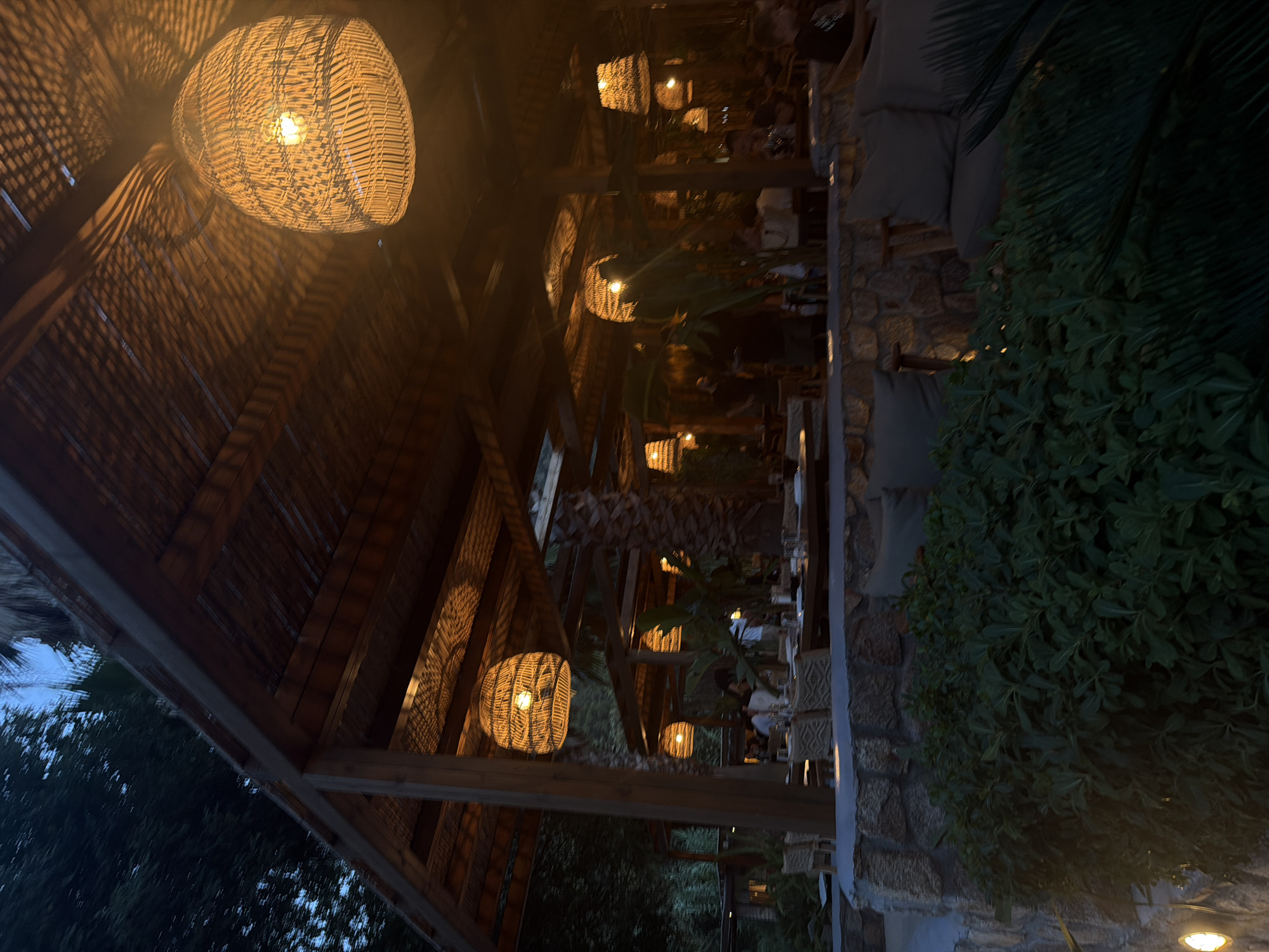 The image shows an outdoor restaurant seating area with a thatched roof. Multiple large, woven pendant lights hang from the ceiling, casting warm, patterned shadows. The seating area features tables set with tableware and chairs. Stone walls and lush green foliage surround the dining space. Several people are visible in the background, seated at tables and moving around the restaurant. The lighting suggests it is evening or dusk. The overall ambiance is relaxed and inviting. This scene is located at a restaurant in the Sithonia Municipal Unit, Greece.