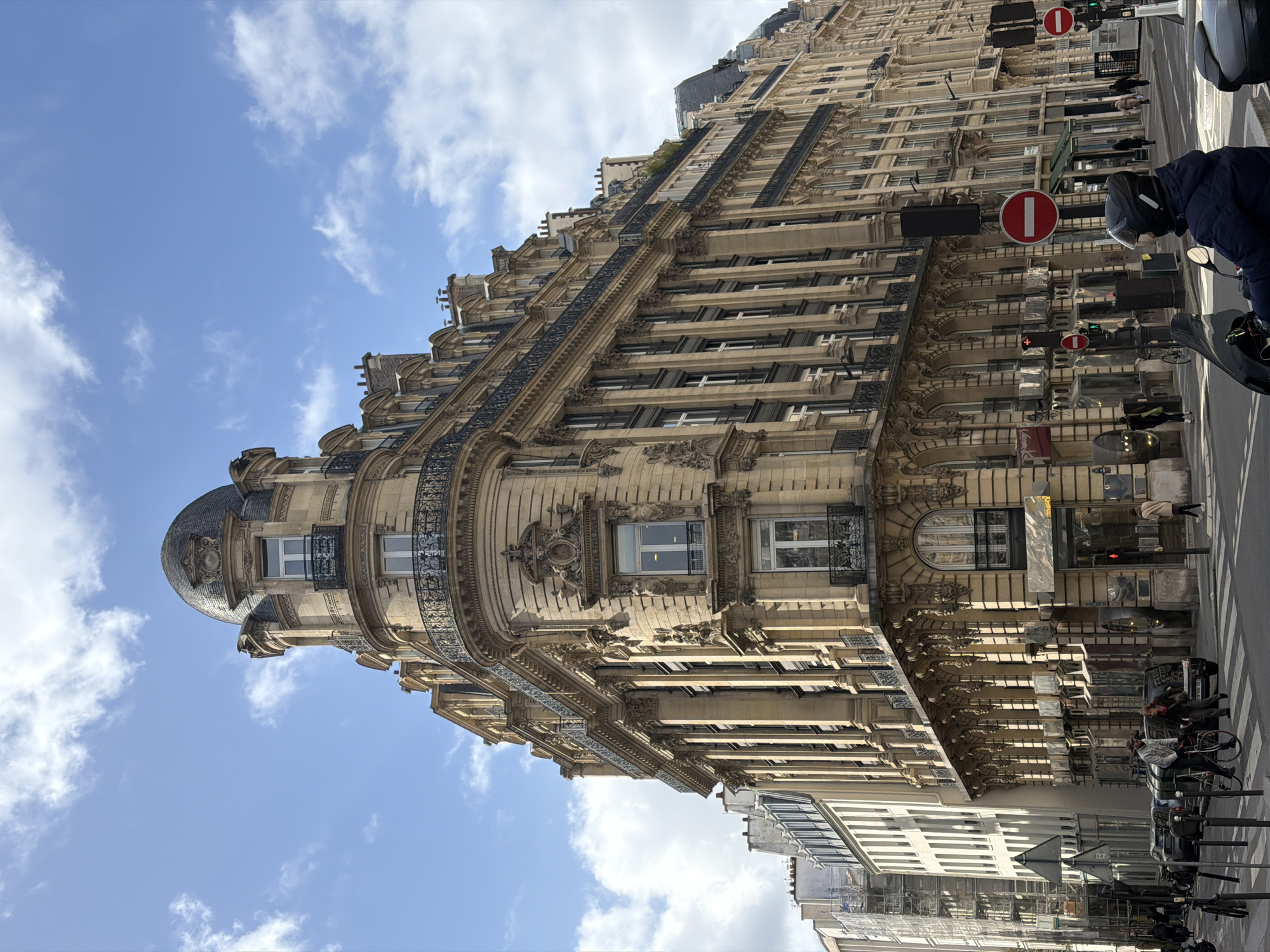 The image shows a tall, ornate building in Paris, France, taken from a street-level perspective. The building features classical architecture with multiple stories, detailed moldings, iron balconies, and a domed turret at its corner. The exterior is primarily light beige in color.
The sky above is blue with scattered white clouds. Part of the street is visible, showing parked bicycles and a person on a scooter wearing a helmet. There are also street signs and traffic lights along the side of the building. A "no entry" traffic sign can be clearly seen. Other buildings are visible in the background.
