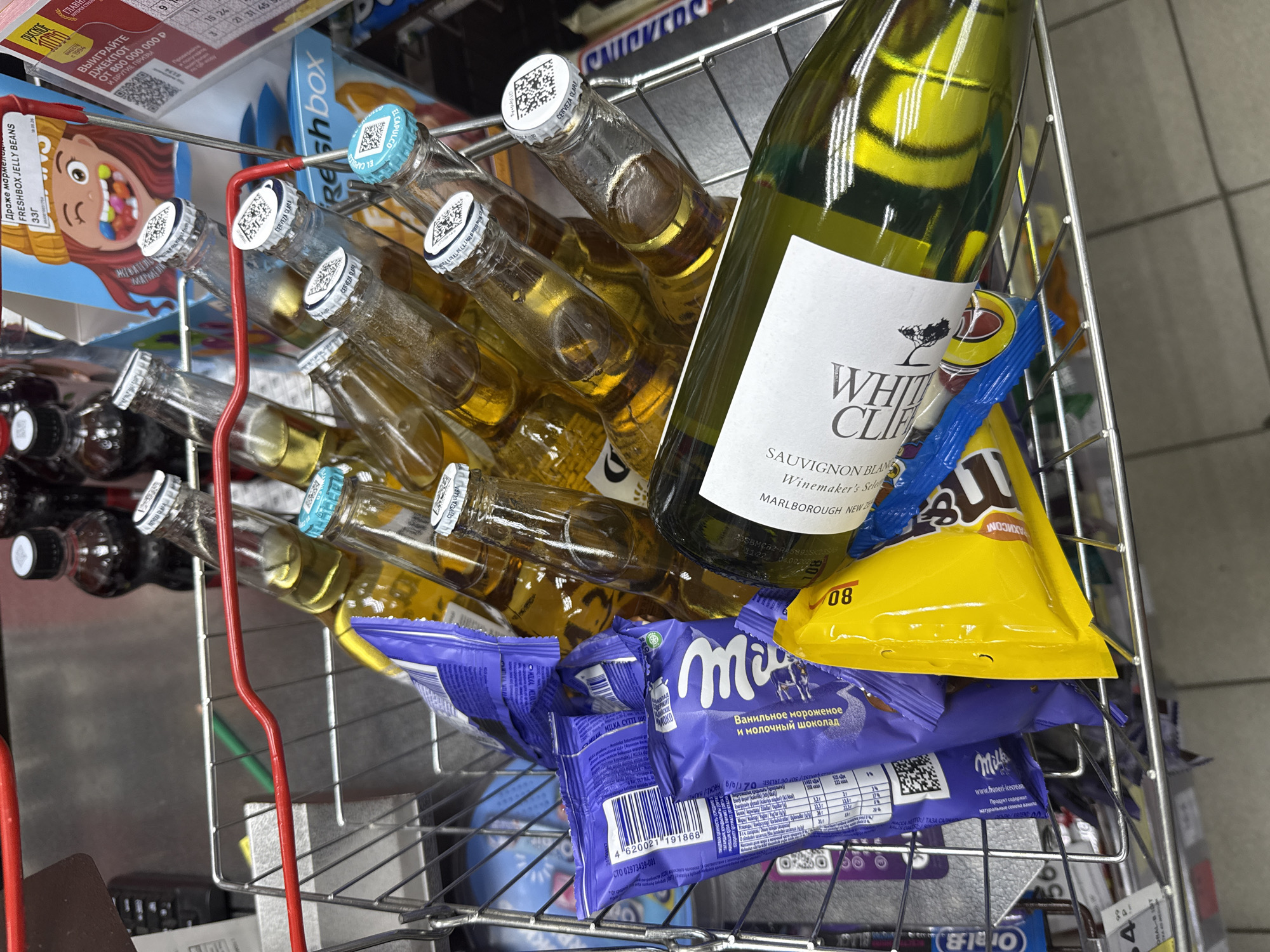 Captured from above, the image showcases a wire shopping basket filled with various items. Dominating the frame are multiple bottles of what appears to be light-colored beer, with caps bearing QR codes. A bottle of white wine labeled "White Cliff Sauvignon Blanc" is also present. Beneath these beverages, there's an assortment of confectionery items, including "Milka" chocolate bars, a yellow bag, and a box labeled "Freshbox Jelly Beans". The basket's red handle is visible on the left. The scene likely occurs in a retail environment in the city of Yemelyanovo, Russia.