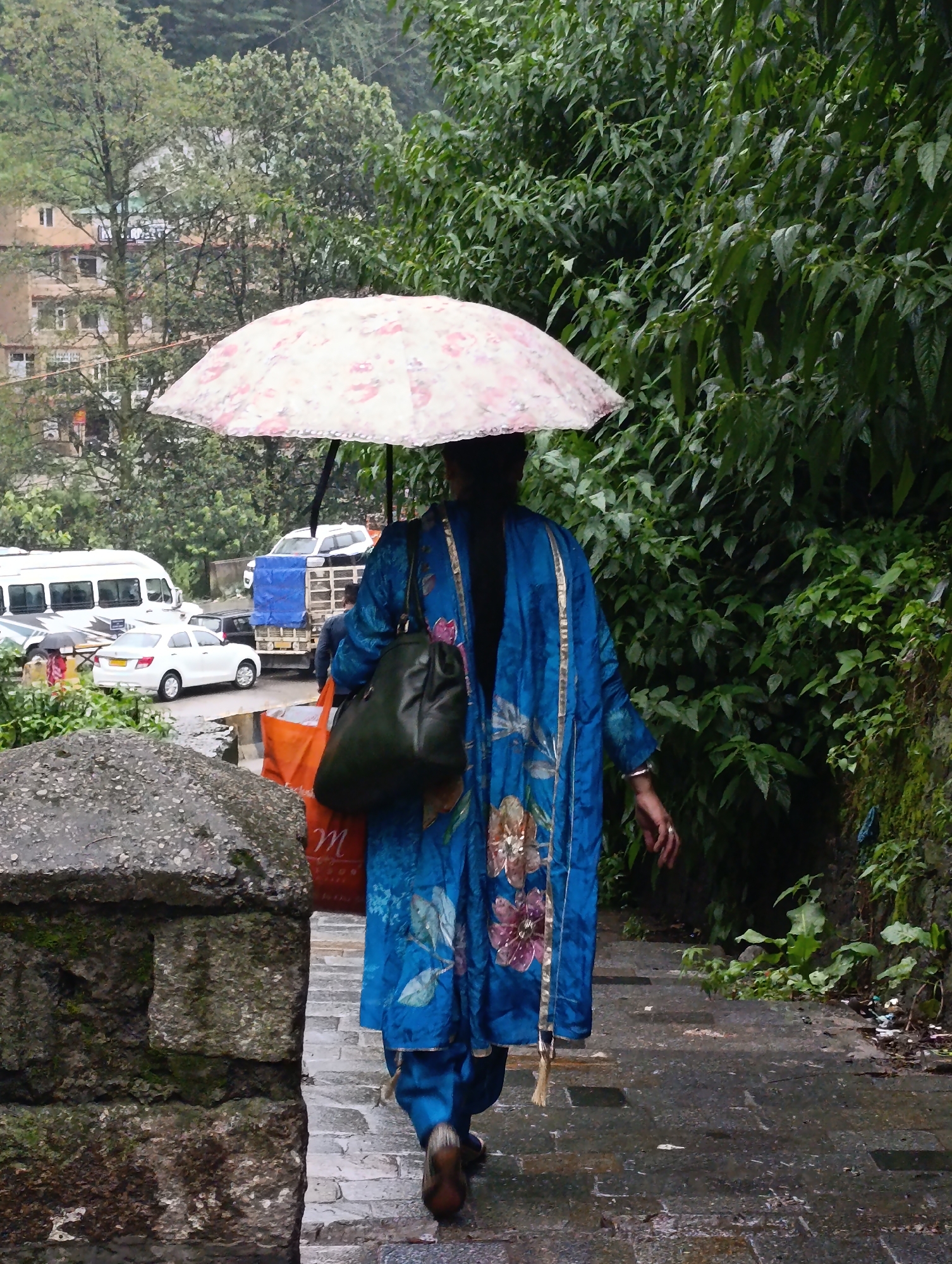 In Manali, India, on a damp stone staircase descending amidst lush foliage, a person walks away from the camera under a large, floral-patterned umbrella. The individual wears a vibrant blue, embroidered robe and dark pants, carrying a dark green handbag on their left shoulder and a bright orange bag in their right hand. The scene suggests a rainy or misty day, with wet stone steps and reflections on the surface. In the background, vehicles, including a white van and a white car, are visible on a road lined with buildings and dense greenery. The atmosphere is one of a pedestrian pathway in a hilly, urban environment, likely a market area or residential street.