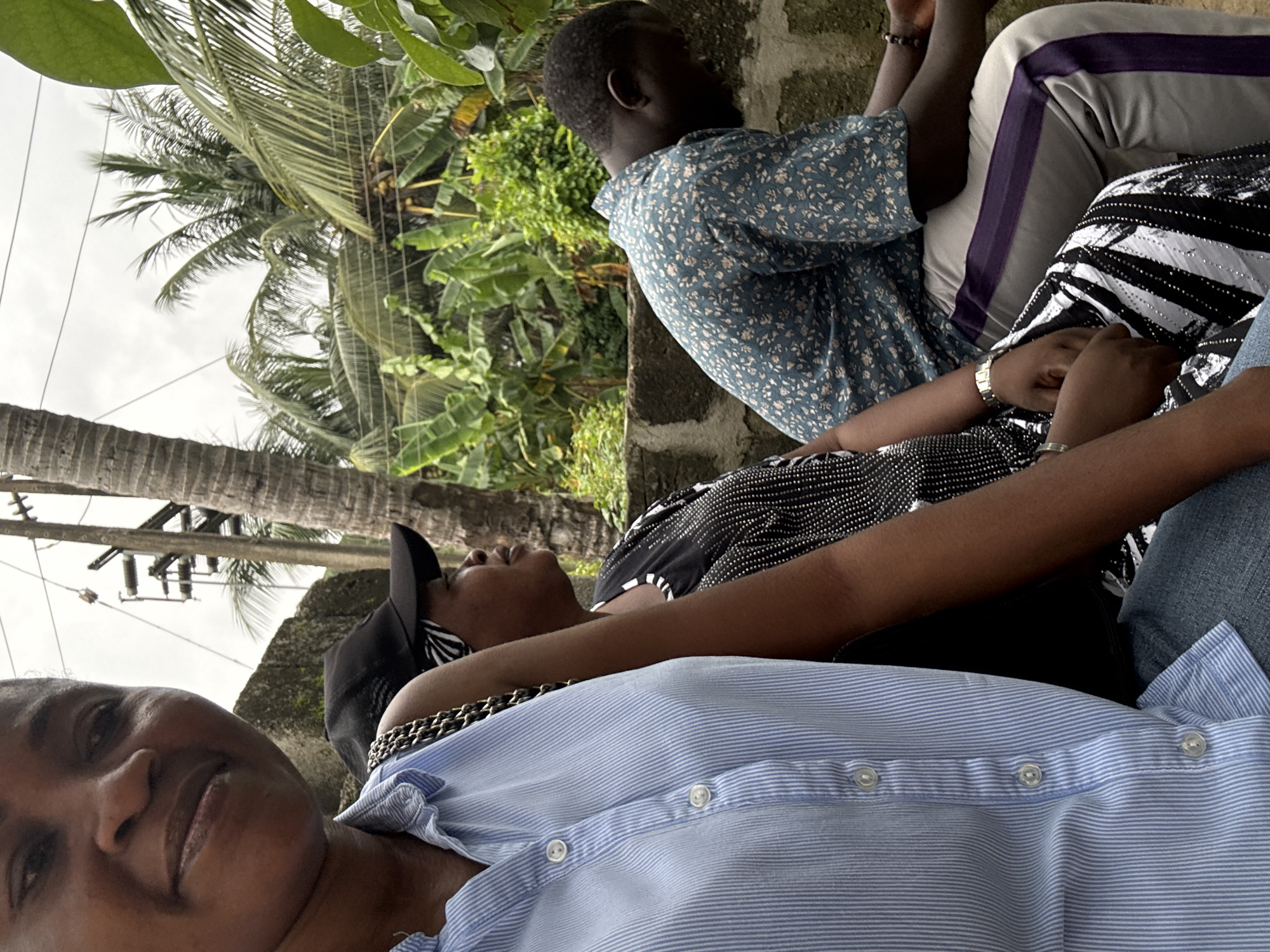This is a vertically oriented photograph of four individuals, likely friends or family, seated together outdoors. The scene is lush and tropical, with prominent palm trees and other green foliage framing the subjects. The sky appears overcast, suggesting it might be a cloudy day or early morning/late afternoon.
In the foreground, a woman in a light blue pinstripe shirt is looking directly at the camera, her expression relaxed and her head tilted slightly. She is wearing a gold-toned chain necklace. To her left and slightly behind her, another person is seen wearing a black and white striped top with shiny embellishments. This individual has their arms resting on their lap.
Behind them, a person in a blue floral print shirt is partially visible, their back to the camera. They appear to be looking to the left, towards the foliage. Further back, a fourth individual is mostly obscured, wearing grey pants with purple and white stripes. Their legs are crossed, and they are also looking away from the camera.
The group is seated on what appears to be a low wall or ledge, possibly part of an outdoor seating area. The overall mood seems casual and relaxed, with the subjects enjoying each other's company in a natural, verdant setting. There is no visible text in the image.