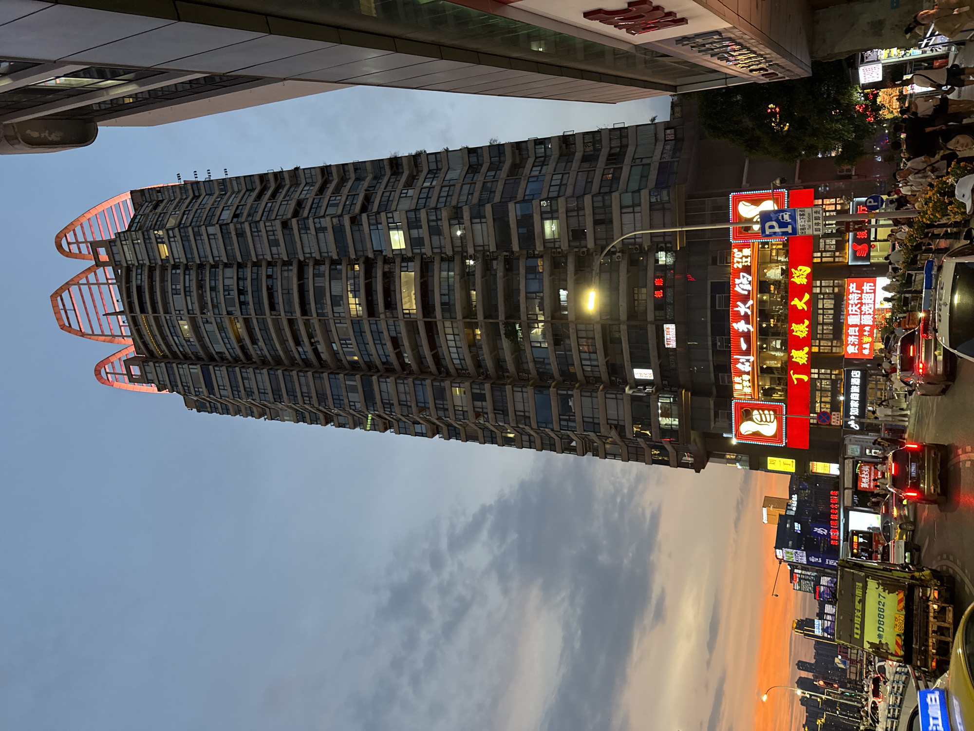 The image captures an urban scene at dusk in Yuzhong District, China. A tall, multi-story building with a distinctive, illuminated crown-like structure on its roof dominates the frame. The building's facade is characterized by numerous windows and balconies, with some lights on, indicating occupancy. Adjacent to this skyscraper, another building with a modern facade is partially visible on the right.
Below the buildings, a street is bustling with traffic, including cars, a garbage truck, and what appears to be a police vehicle. Pedestrians are visible on the sidewalk, some interacting with each other, and others walking along. Numerous illuminated signs with Chinese characters are displayed on the buildings, advertising various businesses, including restaurants and a hotel. The sky is a gradient of blues and oranges, with some clouds present, suggesting the sun has recently set or is about to rise. The overall impression is a vibrant city scene at twilight, with artificial lights illuminating the architecture and streets.