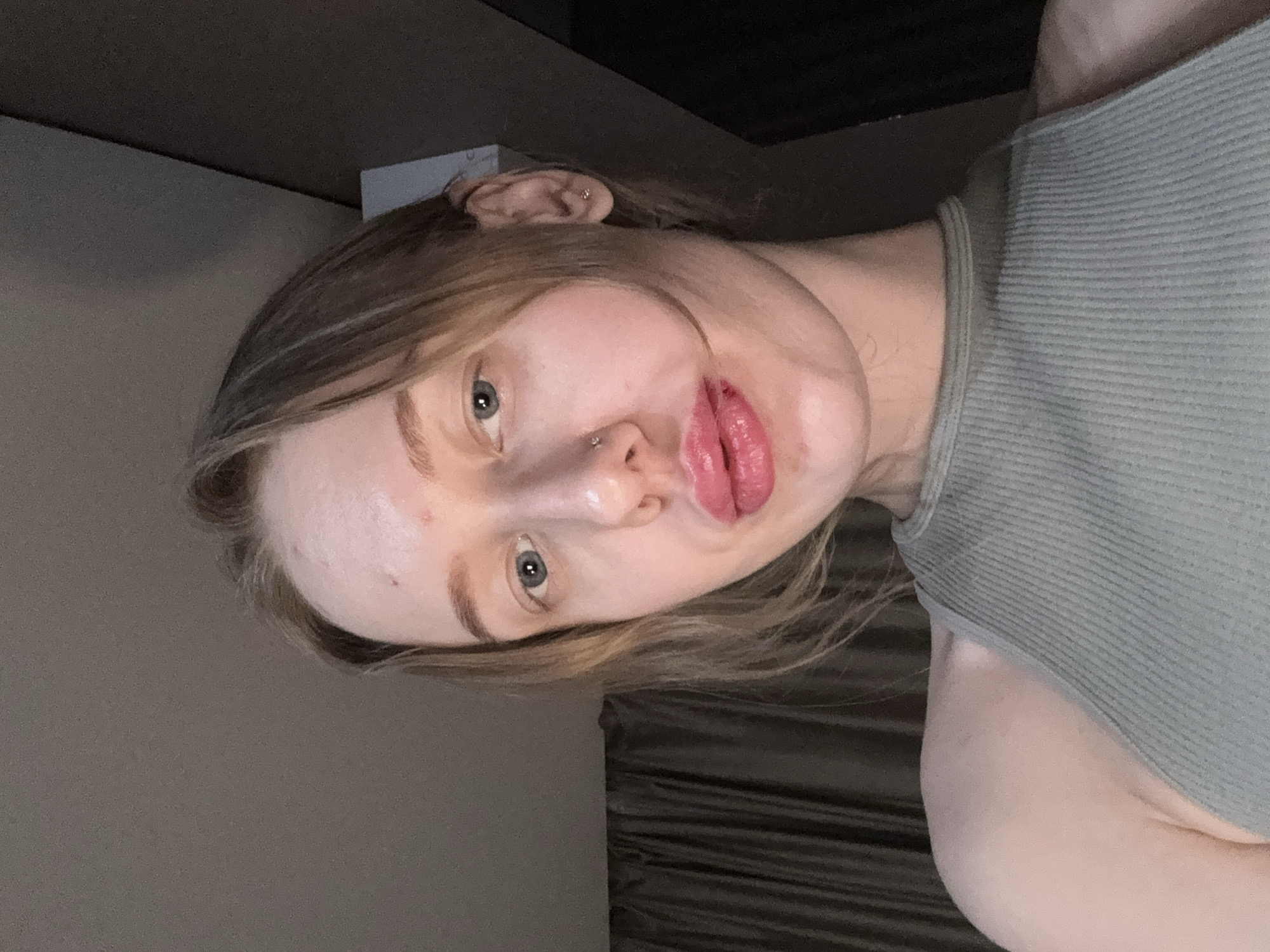 A young woman with fair skin, light brown hair, and blue eyes poses for a selfie. She wears a gray ribbed tank top and has a nose piercing. The background is a dimly lit room with a textured wall and a glimpse of a white object near the top left corner. The location is not discernible as Rostov-on-Don, Russia based on the provided image.