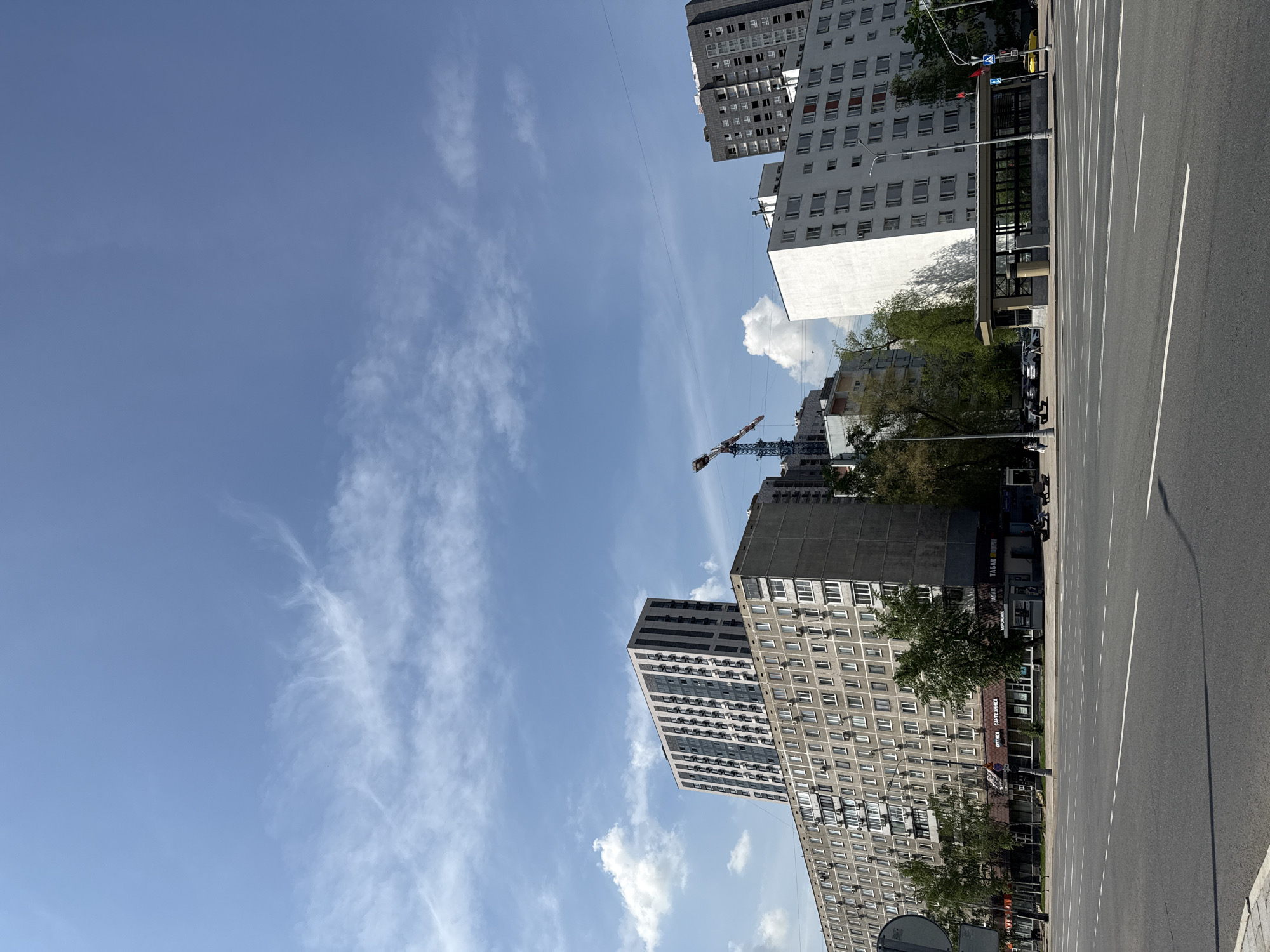 The image shows a street scene in Moscow, Russia, with a view of apartment buildings and a construction crane against a blue sky with scattered clouds. A wide, empty road runs along the right side of the image, marked with white lines. The buildings vary in height and architectural style, ranging from older Soviet-era structures to more modern high-rises. Trees are visible along the street and among the buildings. Shops and other businesses line the base of the apartment buildings.
