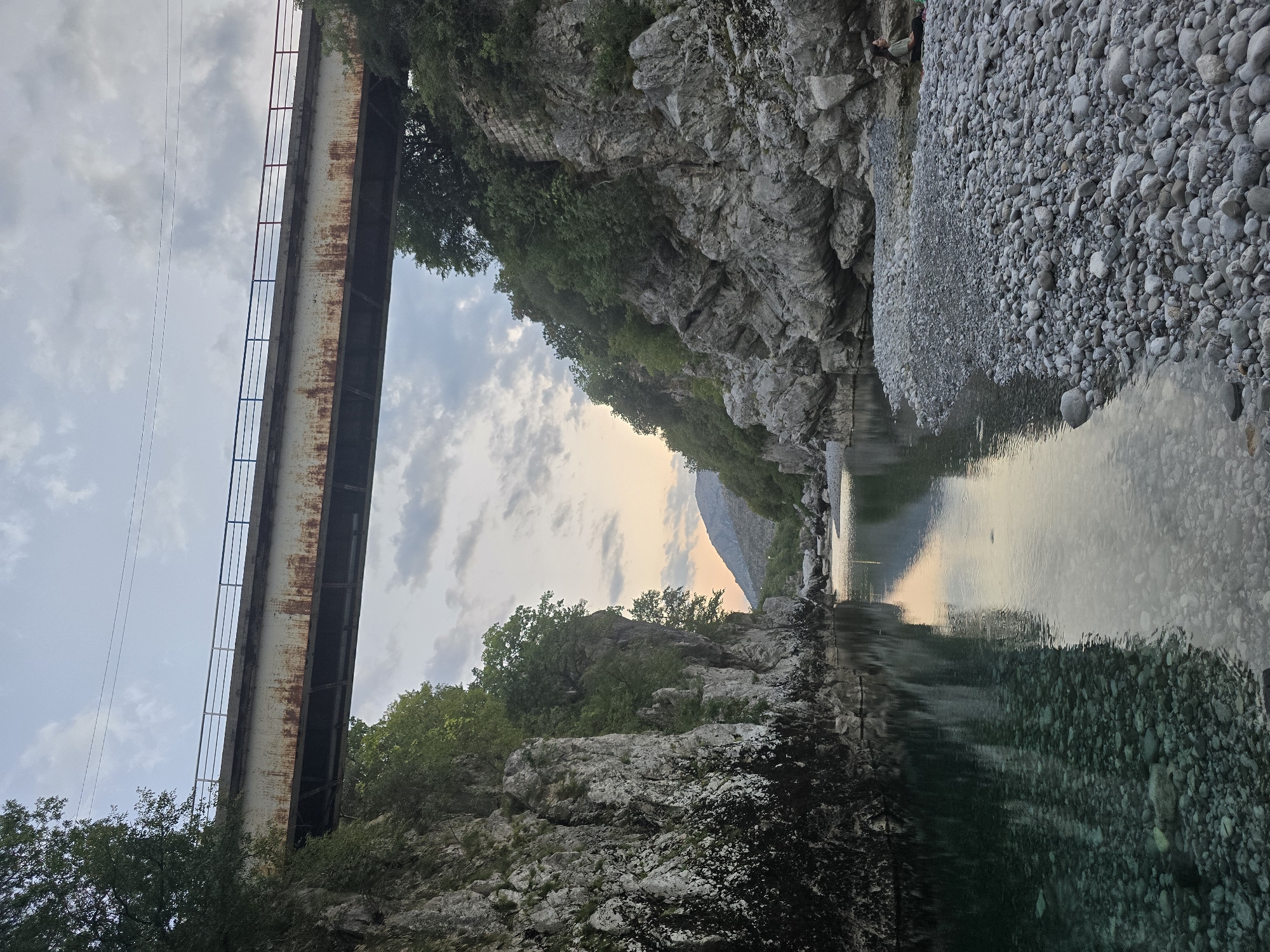 The image depicts a tranquil river flowing through a rocky gorge in Bioče, Montenegro. A sturdy, weathered bridge with rusted beams spans the gorge overhead. The river's clear, shallow waters reveal smooth, rounded pebbles on its bed. Lush green trees cling to the steep, rocky cliffs that line the riverbanks. In the distance, rolling hills and a mountain range are visible under a cloudy sky. A lone figure sits on the pebbled shore to the right, engaged in a moment of quiet contemplation. The scene is illuminated by the soft, ambient light of either dawn or dusk.