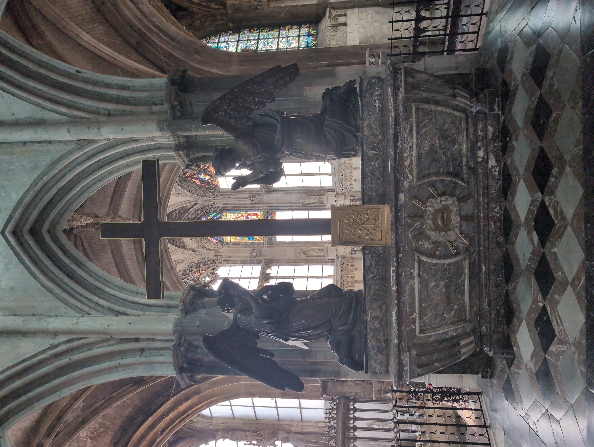 The image depicts the interior of a cathedral, specifically Saint-Jacques Church in Le Tréport, France. Dominating the center of the frame is a large, dark wooden cross on a marble altar. Flanking the cross, kneeling on either side, are two bronze statues of angels with spread wings. The angels are in prayerful poses, with their hands clasped together in front of their faces. The altar itself is a substantial structure, adorned with marble and featuring decorative elements. Behind the altar, large stained-glass windows with intricate geometric patterns and figures are visible, casting colorful light into the space. The architectural style of the cathedral is evident in the vaulted ceilings and arched windows. The floor is covered in a black and white checkered marble pattern. In the background, metal railings and parts of the church’s gothic architecture can be seen. The overall impression is one of solemnity and reverence.