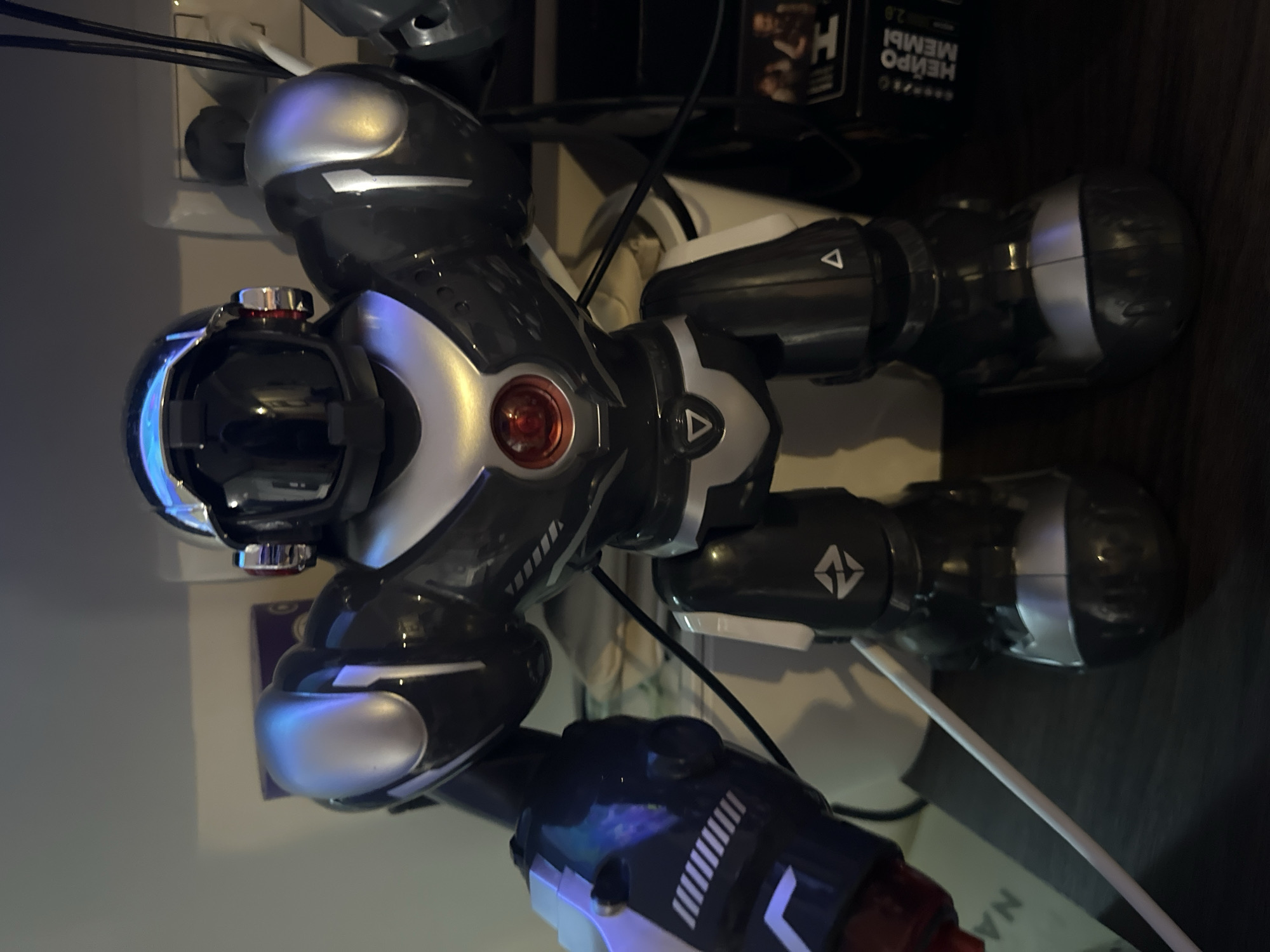A black and silver robot toy is standing on a dark surface, possibly a shelf or desk. The robot has a shiny black body with silver accents on its shoulders, chest, and helmet. It has a red circular light on its chest and a black visor. One of its arms is positioned in front of its body, and appears to have some blue and white detailing. The background is blurred and appears to be an indoor environment with electrical outlets and some boxes on the right side. The lighting is dim, with some reflections on the robot's metallic surfaces. The specific location in Kathu, Thailand is not discernable from the image.