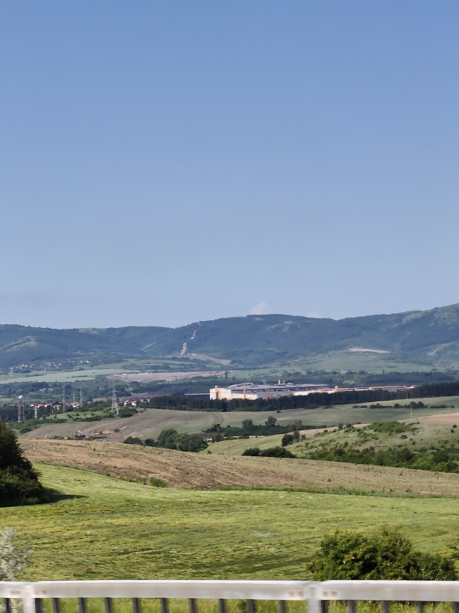 The image shows a panoramic view of a rural landscape with a clear blue sky. In the foreground, there is a white railing. Beyond the railing, rolling green hills and fields stretch into the distance. Several patches of tilled or dry earth are visible among the green fields, indicating agricultural activity.
In the middle ground, a small settlement with red-roofed houses and several tall electricity pylons can be seen. Further back, a large industrial complex with a long, light-colored building dominates the landscape. This complex appears to be situated in a more developed area, contrasting with the surrounding natural scenery.
The background features a range of rolling, tree-covered hills and mountains, with a lighter patch visible on the slope of one of the mountains, possibly indicating a landslide or an area of exposed earth. The scene appears to be a sunny day, with good visibility.
This vista is characteristic of the area around Dragichevo, Bulgaria, known for its combination of agricultural land, industrial zones, and natural mountainous terrain. The image captures a typical view of the Bulgarian countryside with elements of both rural life and industrial development.