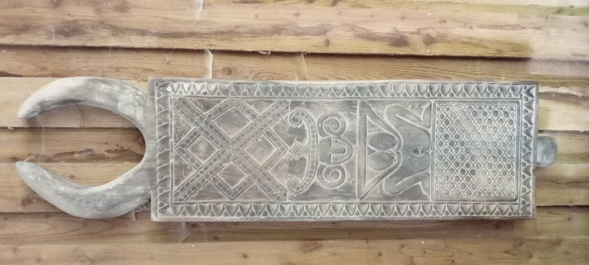 An ornamental wooden carving, resembling a crescent-shaped headdress or horns, is mounted vertically on a wooden plank wall. The carving is grey and shows intricate patterns, including geometric designs and stylized figures. The wall is made of vertical, light-colored wooden planks with visible grain and some cobwebs. The object appears to be an artifact or decorative piece. The image does not depict any specific location in Gelsenkirchen, Germany.