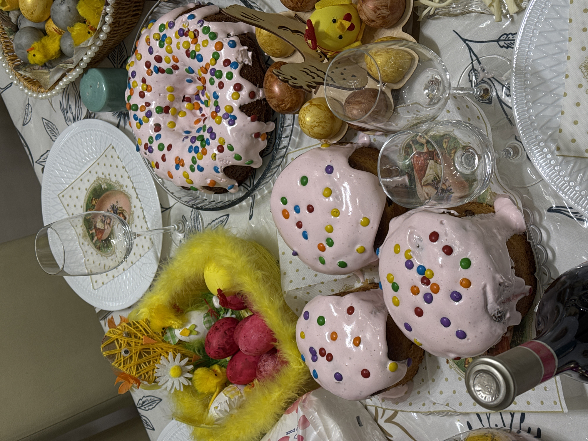 The image shows an overhead view of a table setting, likely for an Easter celebration.
Objects present include:
* Several Easter cakes (Kulich) with pink frosting and candy sprinkles.
* Decorative Easter eggs, some painted gold and brown.
* Small yellow chick decorations.
* A basket filled with Easter decorations.
* Wine glasses.
* A bottle of red wine.
* Decorative plates and napkins.
* A yellow feathered Easter decoration.
The scene suggests a festive gathering with traditional Easter food and decorations. The environment is indoors. There is no information in the image that indicates this scene's location in Yerevan, Armenia.