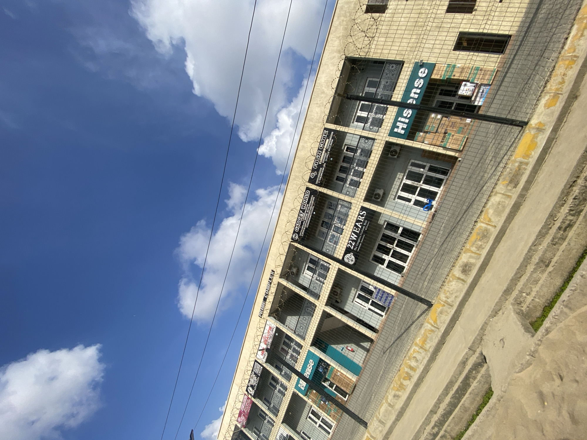 The image shows a multi-story commercial building, possibly a shopping complex, in Ado, Nigeria. The building, labeled "PRIME COMPLEX" along the top, has multiple businesses operating within it, indicated by the signs visible, including "Hi sense", "22 WEARS", "GADGET WORLD", and "FASH CUBE". The architecture features rows of windows, and there is a fence or barrier with what appears to be barbed wire on top along the building's edge. The sky is visible, with clouds against a blue background. The scene is captured from a low-angle perspective. The foreground consists of a sidewalk with steps leading upward. Power lines are visible in the sky.