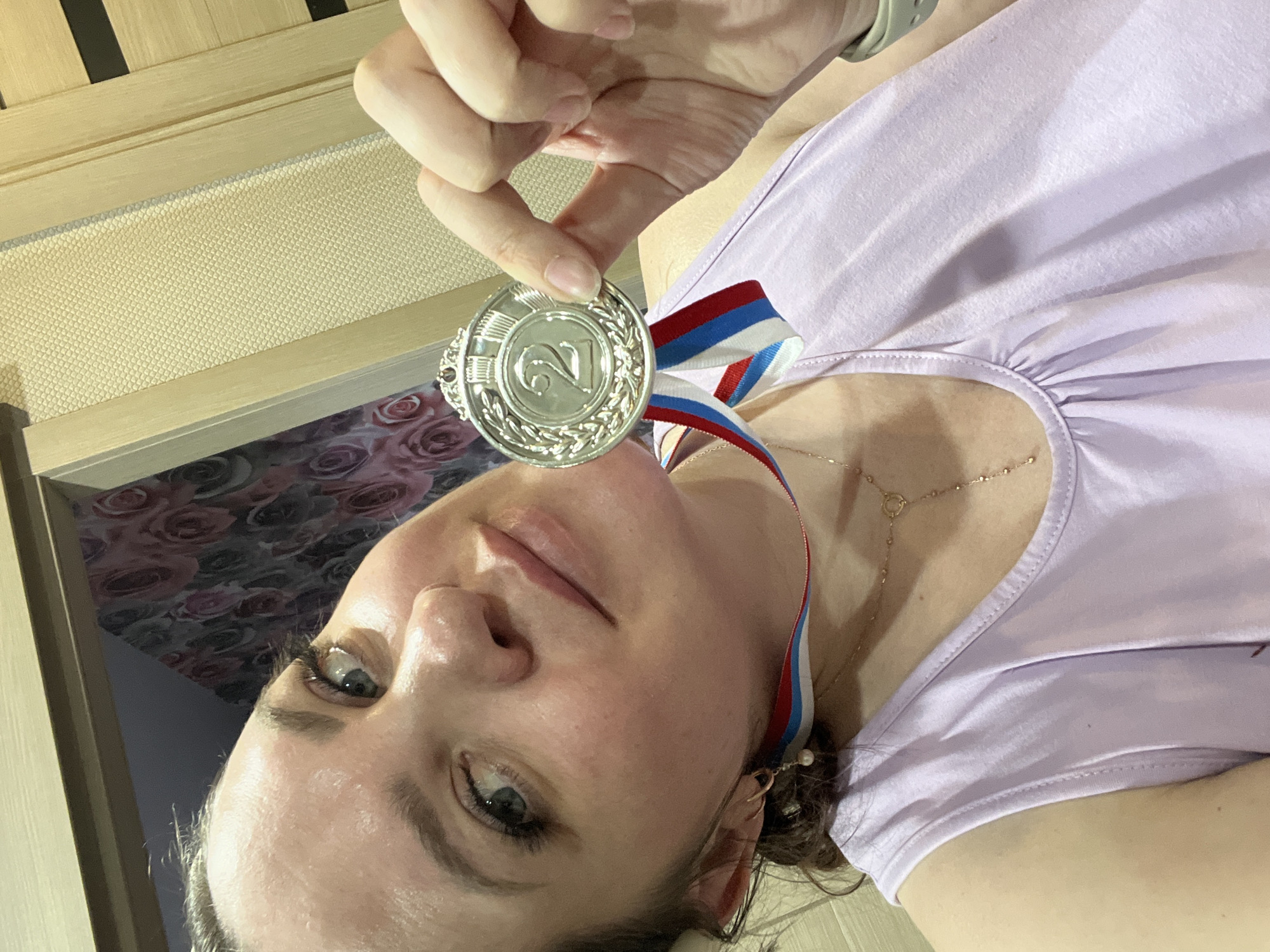 A woman is holding a silver medal with the number "2" on it. The medal has a ribbon with the colors of the Russian flag. She is wearing a lavender t-shirt and a necklace. The background appears to be an interior with a wall. The image has been rotated, and the location Noviy is irrelevant.
