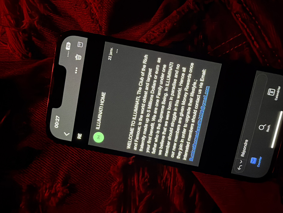 A phone displays a scam email from "ILLUMINATI HOME," promising wealth and a life free of struggle. The message claims membership in the Illuminati, described as the world's oldest and largest fraternity, offers a $5 million welcome bonus. It encourages interested individuals to contact them via email at illuminatihomeofwealth2039@gmail.com. The phone's screen shows the email interface, including reply and search icons. The background is a red, textured surface.
