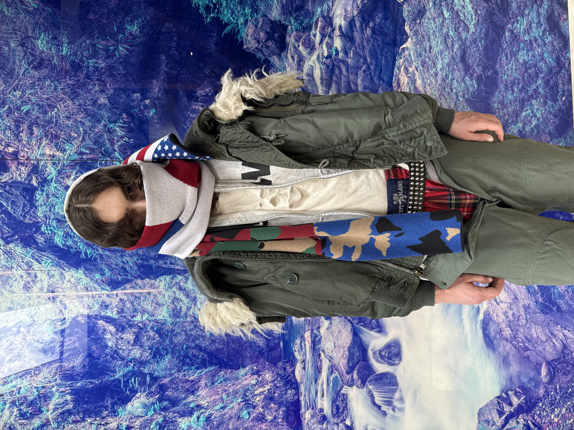 A person stands in front of a wall decorated with a blue and white mountain and waterfall scene. The person is bundled in layers of clothing including a green jacket with a fur-lined hood, a gray sweatshirt with a "W" on the front, a white t-shirt, and a red and black plaid flannel shirt. They are also wearing a studded belt, camouflage and American flag scarves, and olive-colored pants. The setting appears to be indoors, but the exact location and city are not specified.
