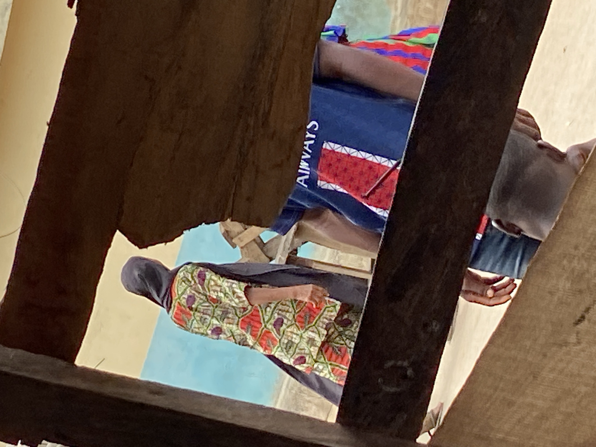 The image offers a downward view into an indoor or semi-enclosed space, framed by dark wooden beams or planks in the foreground, suggesting a perspective from above or through a structural opening.
Three individuals are visible within the brightly lit scene, indicative of daytime. On the left, a person is seated, wearing a vibrant, patterned long dress featuring green, red, and purple motifs, complemented by a dark head covering. Their arm rests on their lap.
In the center-right, another individual is seen from the torso upwards, clad in a dark blue shirt. The white text "ALWAYS" is printed vertically along the side of the shirt, adjacent to a distinct red and white geometric pattern. This person appears to be seated or reclining. Further to the right, partially obscured by a wooden beam, is the head and upper body of a third person, wearing a light-colored head covering, possibly grey, and a blue garment.
The background features light-colored walls or floor surfaces, with sections of light blue and beige/tan. Glimpses of what appear to be simple wooden chairs or stools are visible beneath the individuals. The setting aligns with the provided context of Mararaba, Nigeria, portraying a casual gathering or resting area. The only visible text is "ALWAYS" on the blue shirt.