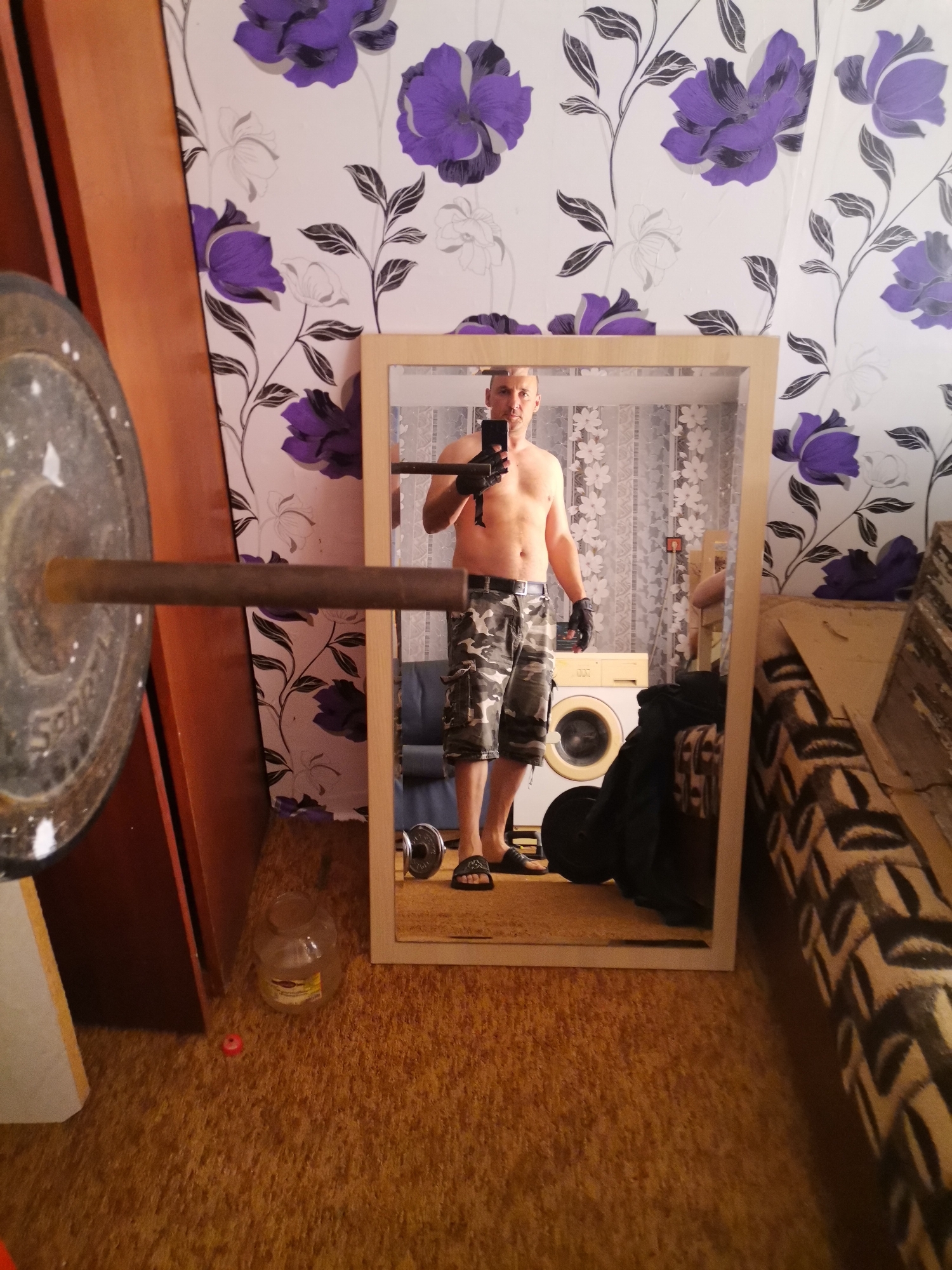 A shirtless man wearing camouflage shorts, black gloves, and sandals is taking a photo of himself in a mirror. The mirror is reflecting a washing machine, a couch with a patterned cover, and floral wallpaper. A barbell with a weight plate is visible in the foreground on the left. A jar and a small red object are on the floor in the lower left corner. The location is unknown.