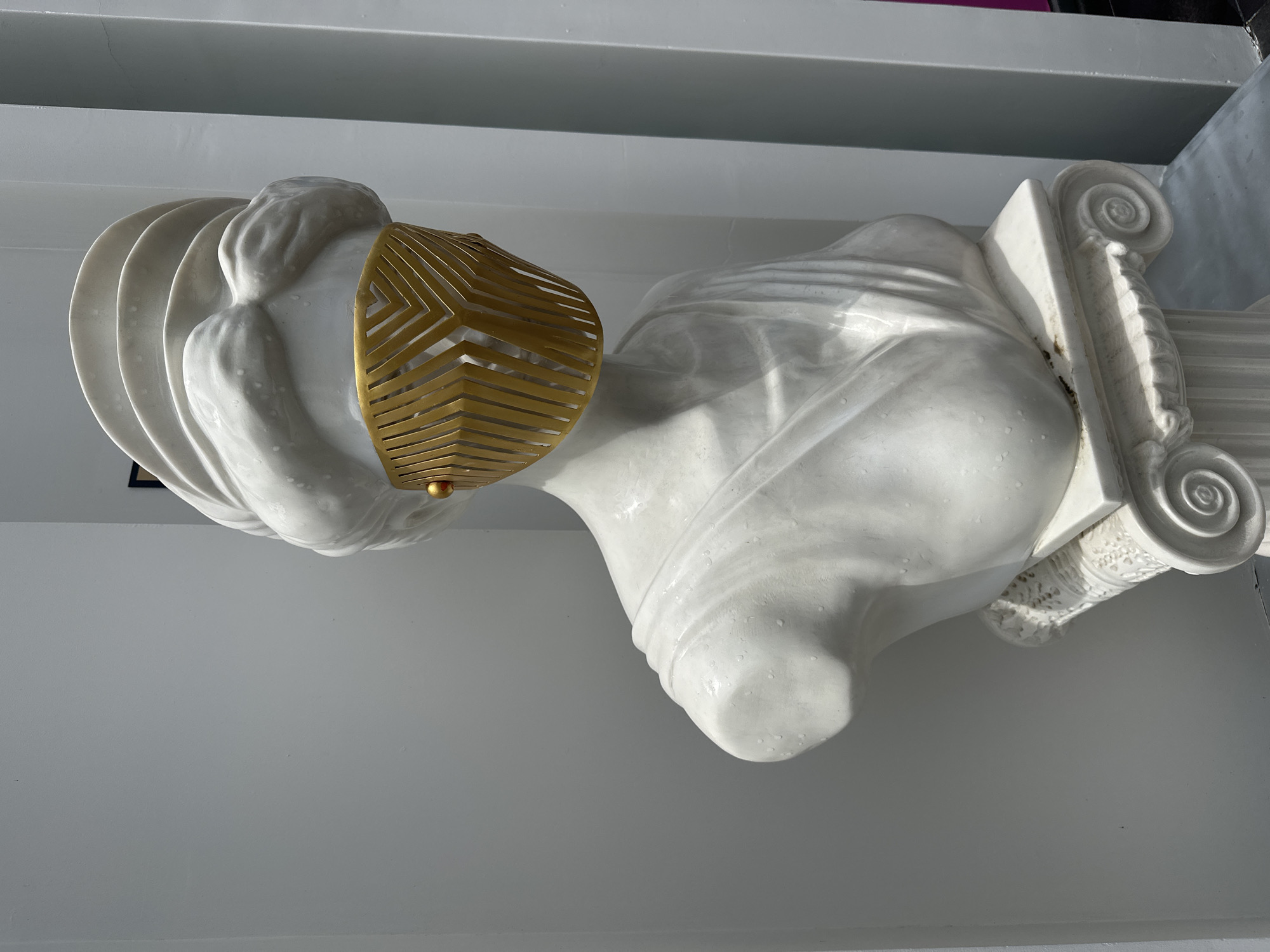 A marble sculpture of a bust of a woman with a gold mask is displayed on a white pillar against a white wall. The statue has traditional Greek or Roman features with draped clothing and ornate details on the pillar. The gold mask obscures the face of the bust, adding a modern element. The environment appears to be indoors. The scene may be in Nha Trang Ward, Bac Nha Trang City, Vietnam.