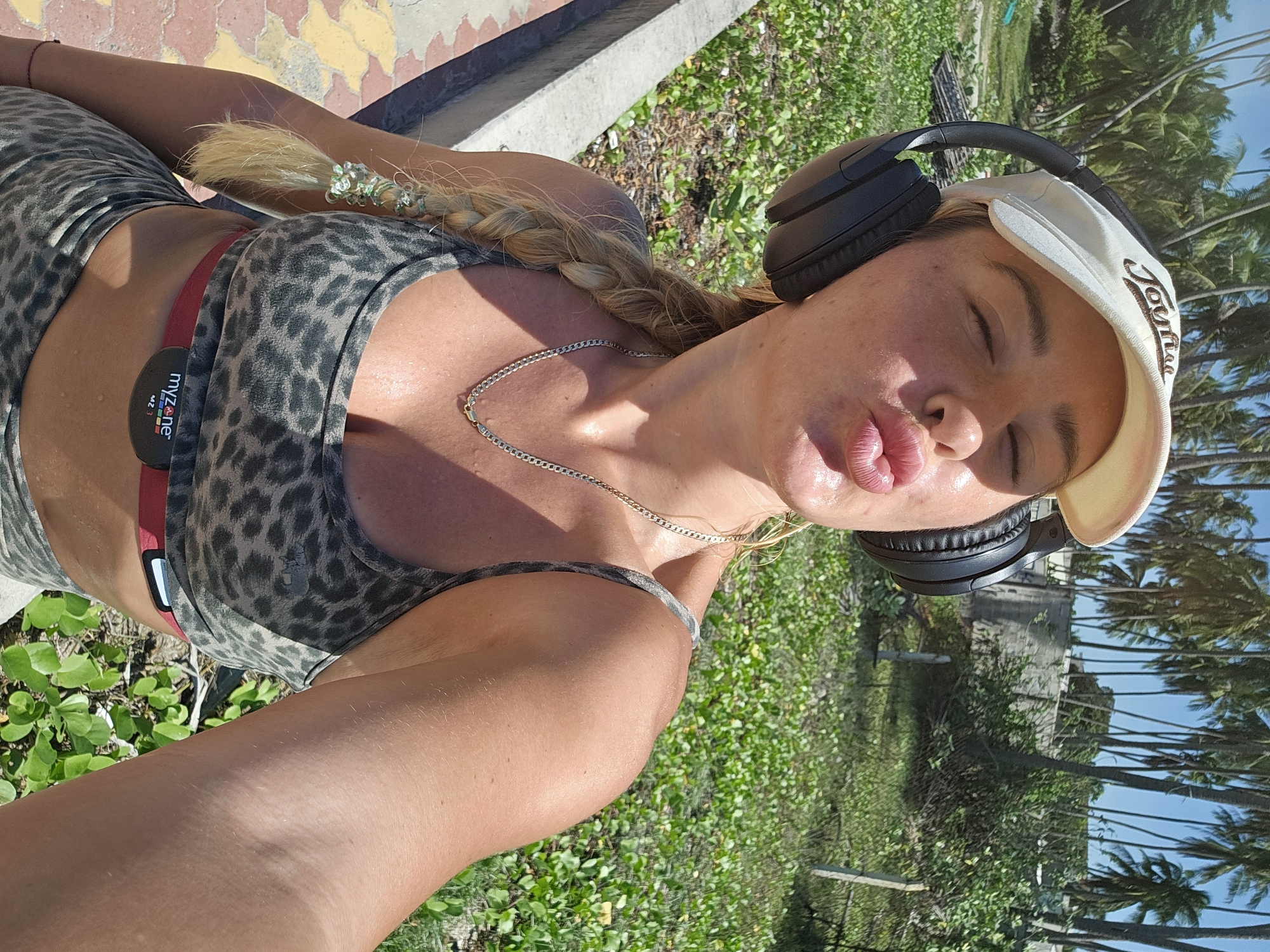 An outdoor, close-up, selfie-style photograph shows a Caucasian woman with blonde hair in a braid. She is wearing a white baseball cap, large black headphones, a leopard-print sports bra, and matching leggings. The woman is making a kissing face with her eyes closed. She is wearing a silver chain necklace and a red MyZone heart rate monitor around her waist. Her skin appears to be sweaty. The background is filled with lush green vegetation and numerous palm trees, suggesting a tropical or subtropical environment. The ground in the foreground is a paved walkway with yellow and red tiles. A concrete barrier runs along the left side of the walkway. The overall impression is that the woman is outdoors, possibly exercising, in a warm climate. The context of Phu Thuy Ward, Vietnam, if this image is indeed from there, would imply a coastal or beachside location given the vegetation and palm trees.