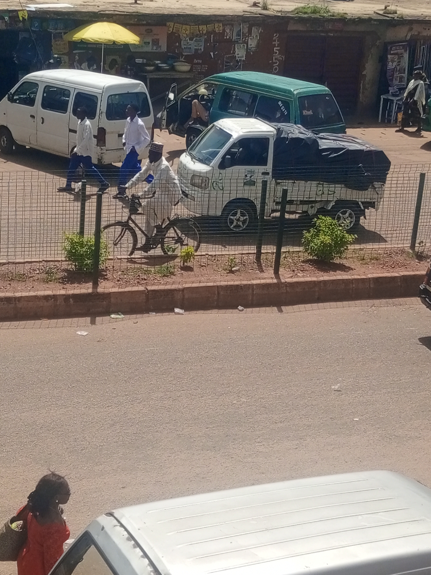 The image depicts a street scene in Bukuru, Nigeria. Several vehicles are visible, including a white minivan, a green van, and a white pickup truck covered with a black tarp. In the foreground, a person is riding a bicycle. Two individuals in white shirts and blue pants are walking, and another person in a white garment and head covering is standing. A woman in a red dress and carrying a bag is on the side of the road in the lower left corner. The scene appears to be set during the daytime, under what seems to be clear weather. The background shows buildings and a yellow umbrella, suggesting a market or roadside stalls. The presence of multiple vehicles and pedestrians indicates a busy street environment.