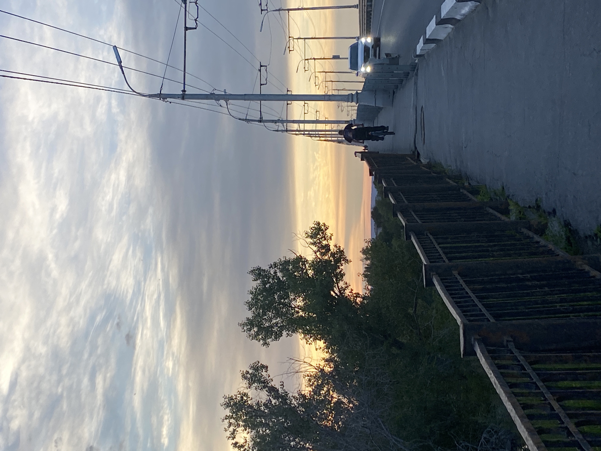 The image depicts a street scene with a person on a bicycle riding on a path next to a road. The setting appears to be a bridge or overpass, indicated by the presence of streetlights and a vehicle driving on the road. A metal railing lines the side of the path, with trees and foliage visible below. The sky is filled with clouds and a sunset is occurring, casting a warm orange and yellow glow. The time of day is clearly sunset, and the weather appears to be clear despite the cloudy sky. The location is Novokuybyshevsk, Russia. There is no visible text in the image.