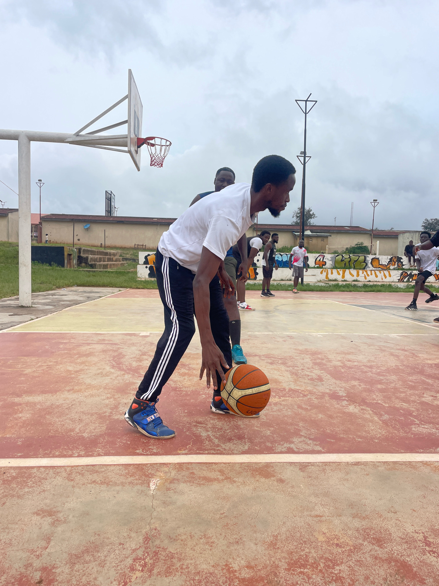 The image captures a group of individuals playing basketball on an outdoor court under an overcast sky. The court surface is a mix of red and yellow concrete with white lines. In the foreground, a man in a white t-shirt and black track pants with white stripes is dribbling an orange basketball, his body low in a defensive stance. He is wearing blue and orange basketball shoes. Behind him, another person in a white t-shirt and dark shorts is visible, also engaged in the game.
Further back on the court, several other individuals are present, some watching and others participating. One person in a white t-shirt and grey shorts stands near a graffiti-covered wall. The background features a building with a red roof, a chain-link fence, and trees. A basketball hoop with a white backboard and a red net is prominently visible on the left side of the frame. The general environment suggests an urban or semi-urban setting. The image does not provide explicit information about the specific location within Gura Top, Nigeria.