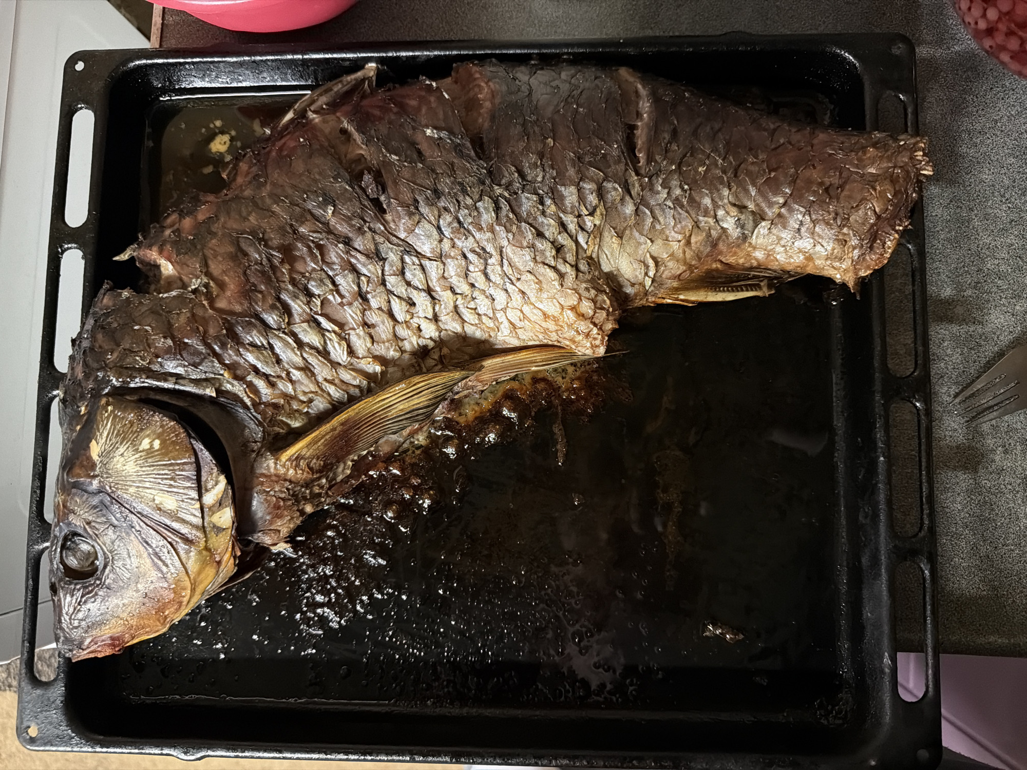 A whole cooked fish, likely carp, lies on a black baking tray filled with dark drippings. The fish is brown and its scales are visible. The tray is placed on a countertop, and part of a pink bowl and a fork are visible in the background. No specific evidence connects this image to Ol'khovo, Russia.