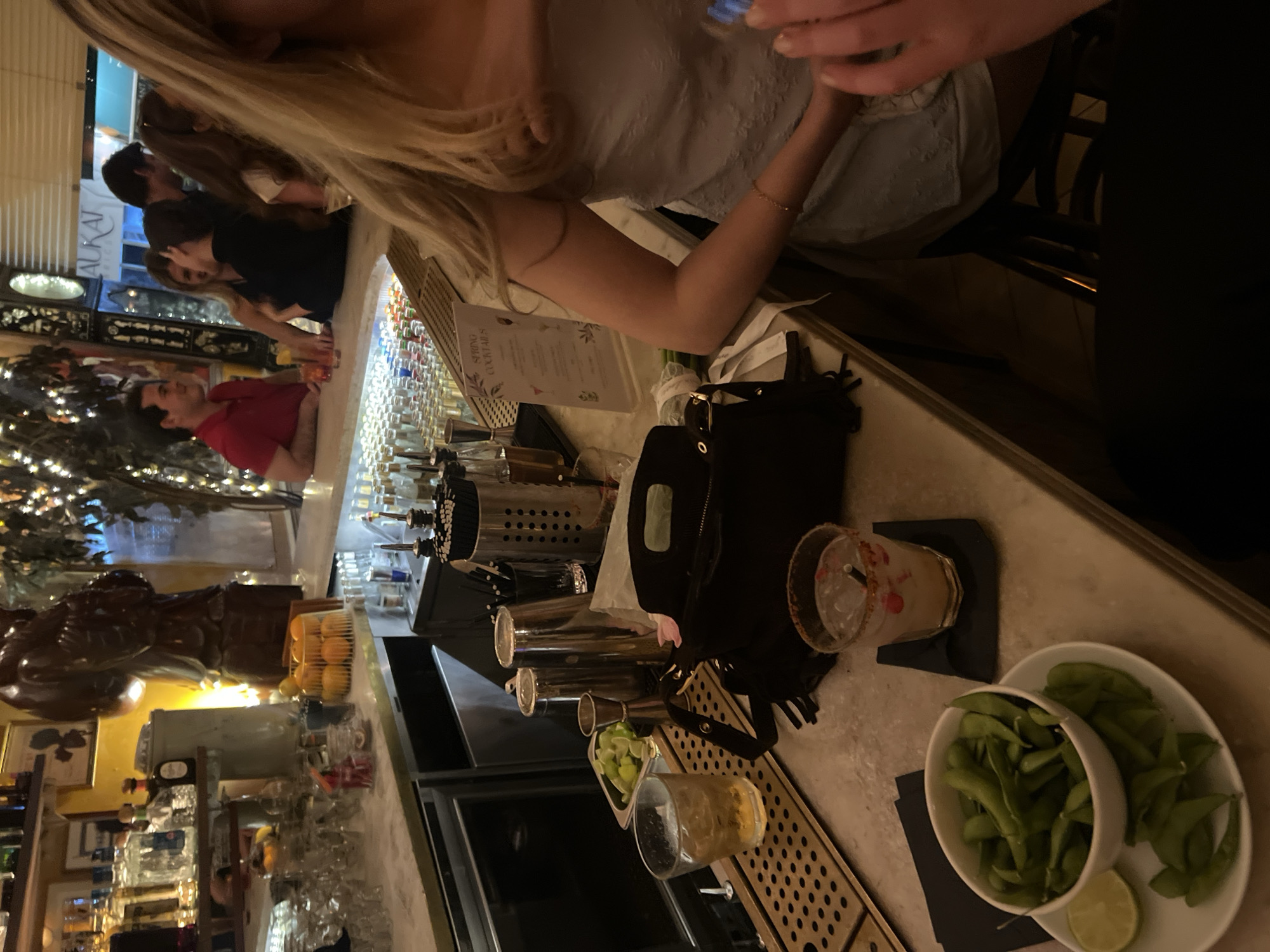 A warmly lit bar scene in London, United Kingdom, shows a group of people gathered at a high bar counter. A woman with blonde hair wears a light-colored dress and sits at the bar next to another person. The bar is stocked with numerous bottles of alcohol and cocktail ingredients. A cocktail menu is visible on the bar counter. A handbag rests on the bar counter, next to a cocktail and a bowl of edamame. A tree decorated with lights appears in the background.