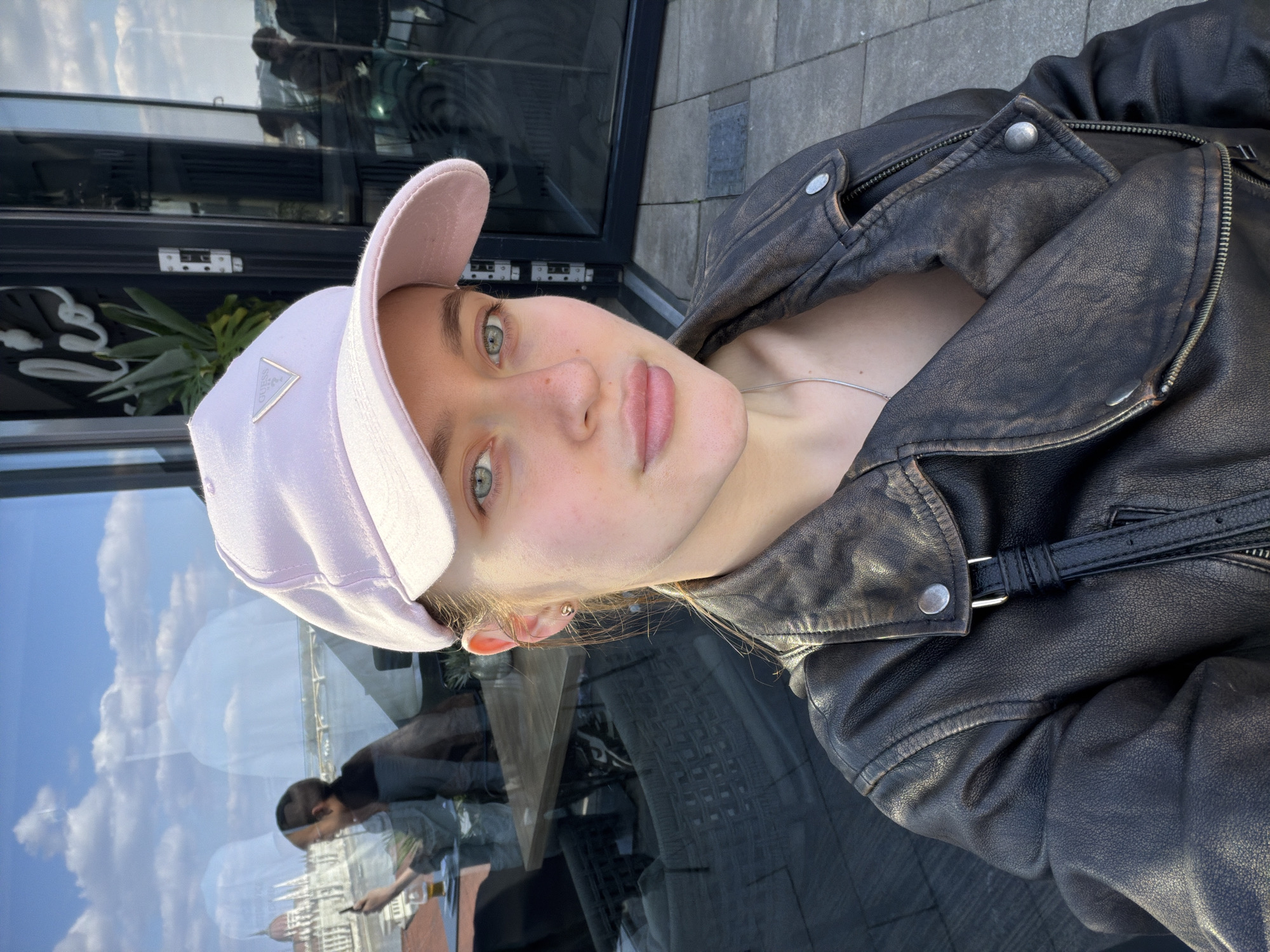 A woman in a pink baseball cap and black leather jacket is photographed. She is near a window, and the reflection shows a man standing with a beverage, with an out-of-focus city landscape including a notable church spire in the background. The location is unknown.