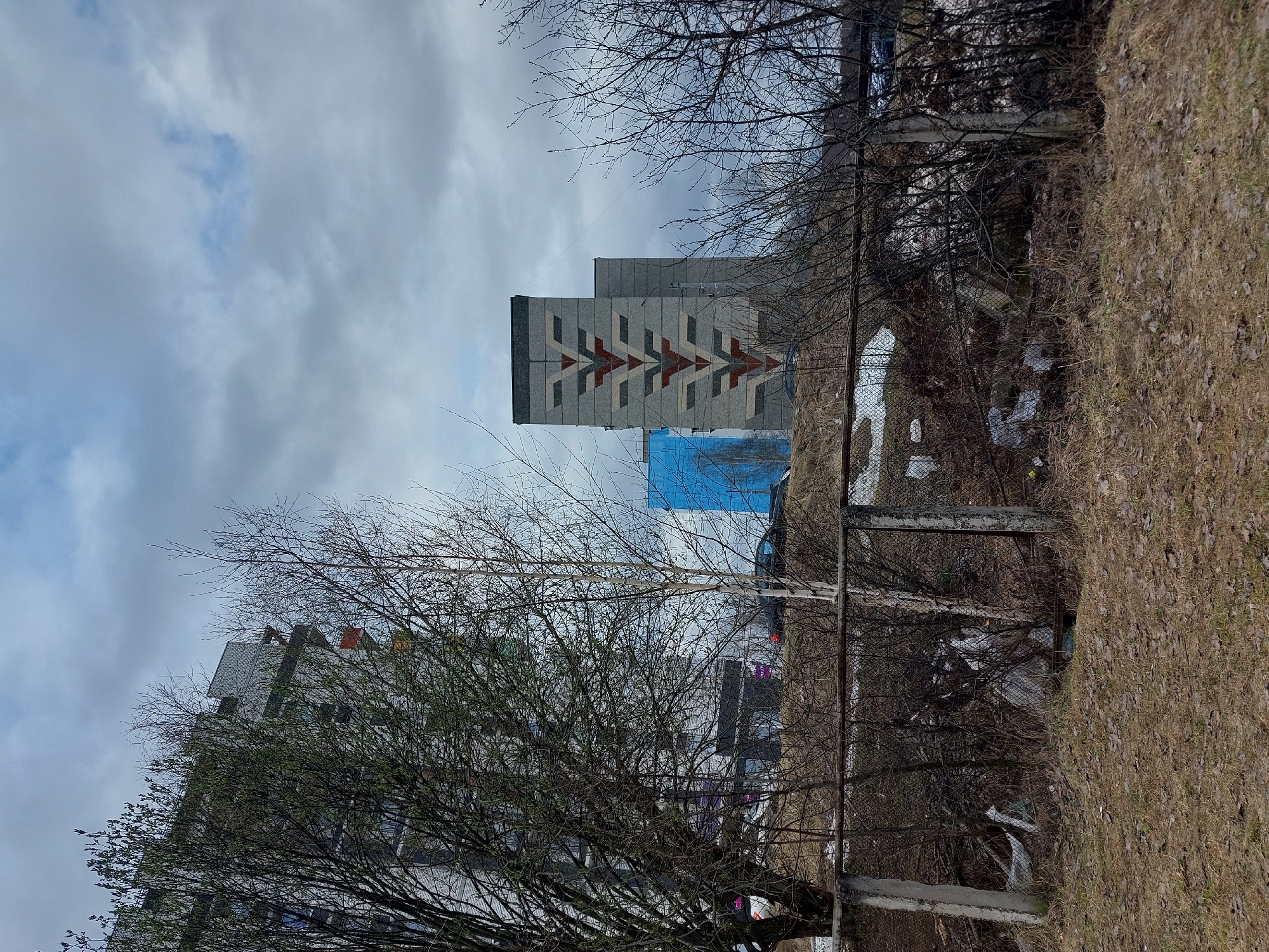 The image shows a gray apartment building in Chaikovsky, Russia. The building has a distinctive chevron pattern in red, gray, and white on its facade. The sky is overcast with gray clouds. A chain-link fence runs along a grassy area in the foreground. Bare trees and branches obscure parts of the building and the surrounding environment. There is some debris and remnants of snow on the ground. A blue tarp is visible covering a section of the building or nearby structure.