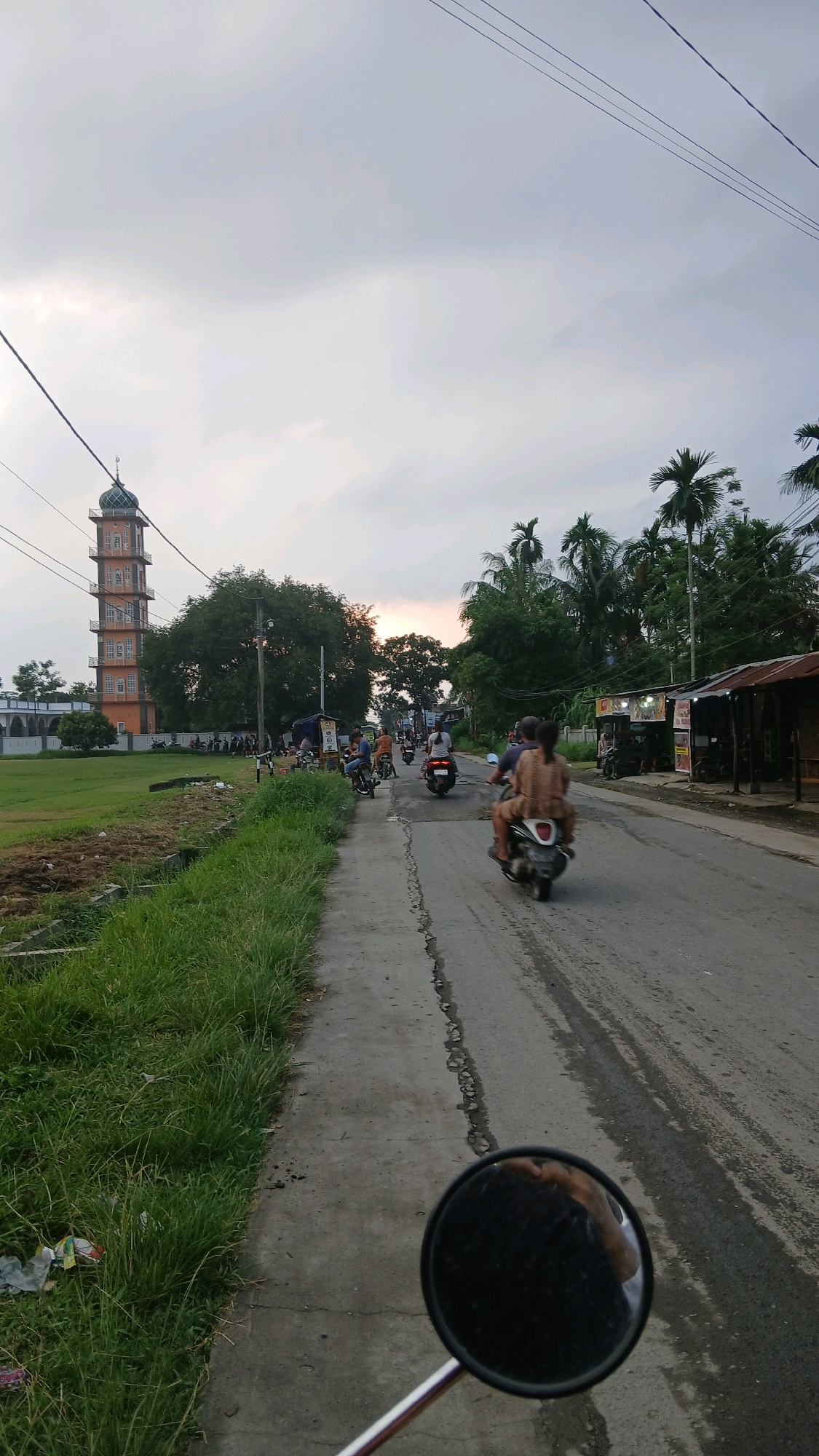 The image depicts a road scene in Sunggal, Indonesia, at dusk. A tall, orange mosque tower with a green dome stands prominently to the left, adjacent to a grassy field and a building. Several people are on motorcycles, with some riding as passengers, moving along the paved road. On the right side of the road are small businesses with corrugated roofs. Lush green grass lines the left side of the road, with some litter visible. The sky is overcast with hints of sunset. The perspective suggests the photo was taken from a moving motorcycle, as a rearview mirror is visible in the foreground.