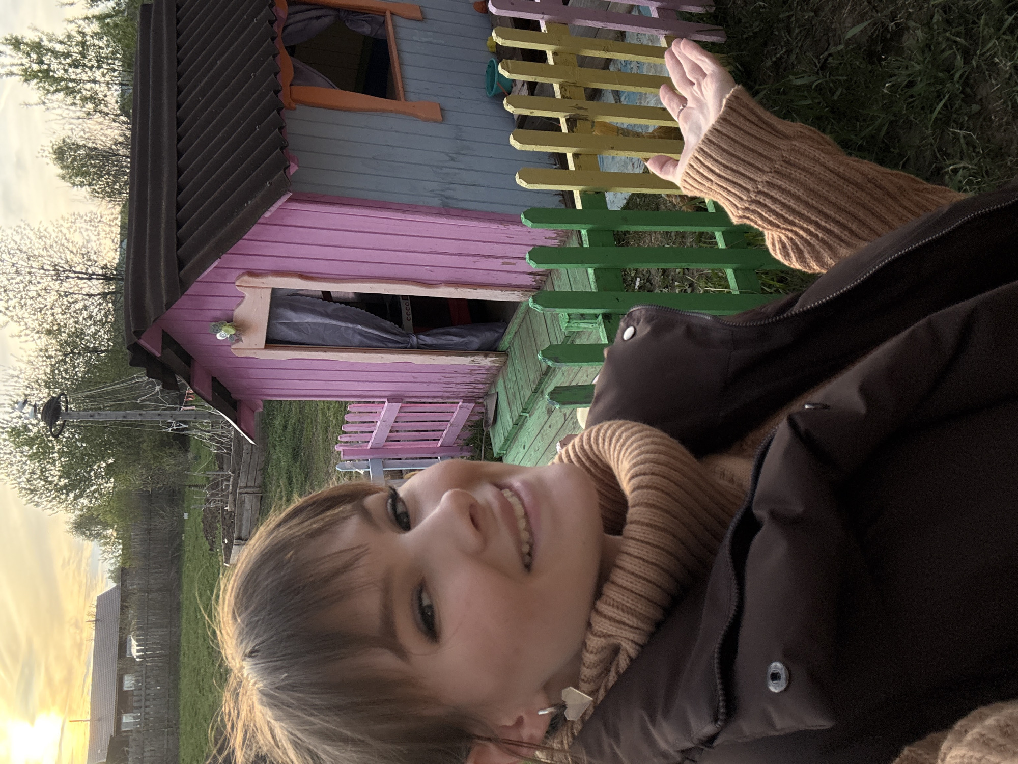 A young woman is lying outdoors, possibly in Курашим, Russia, given the context of the request. She is wearing a brown turtleneck sweater and a dark brown jacket. She is smiling at the camera. Behind her is a small, colorful playhouse with a gray roof, pink sides, and a green and yellow picket fence. In the background, there are trees and a fence visible at sunset.