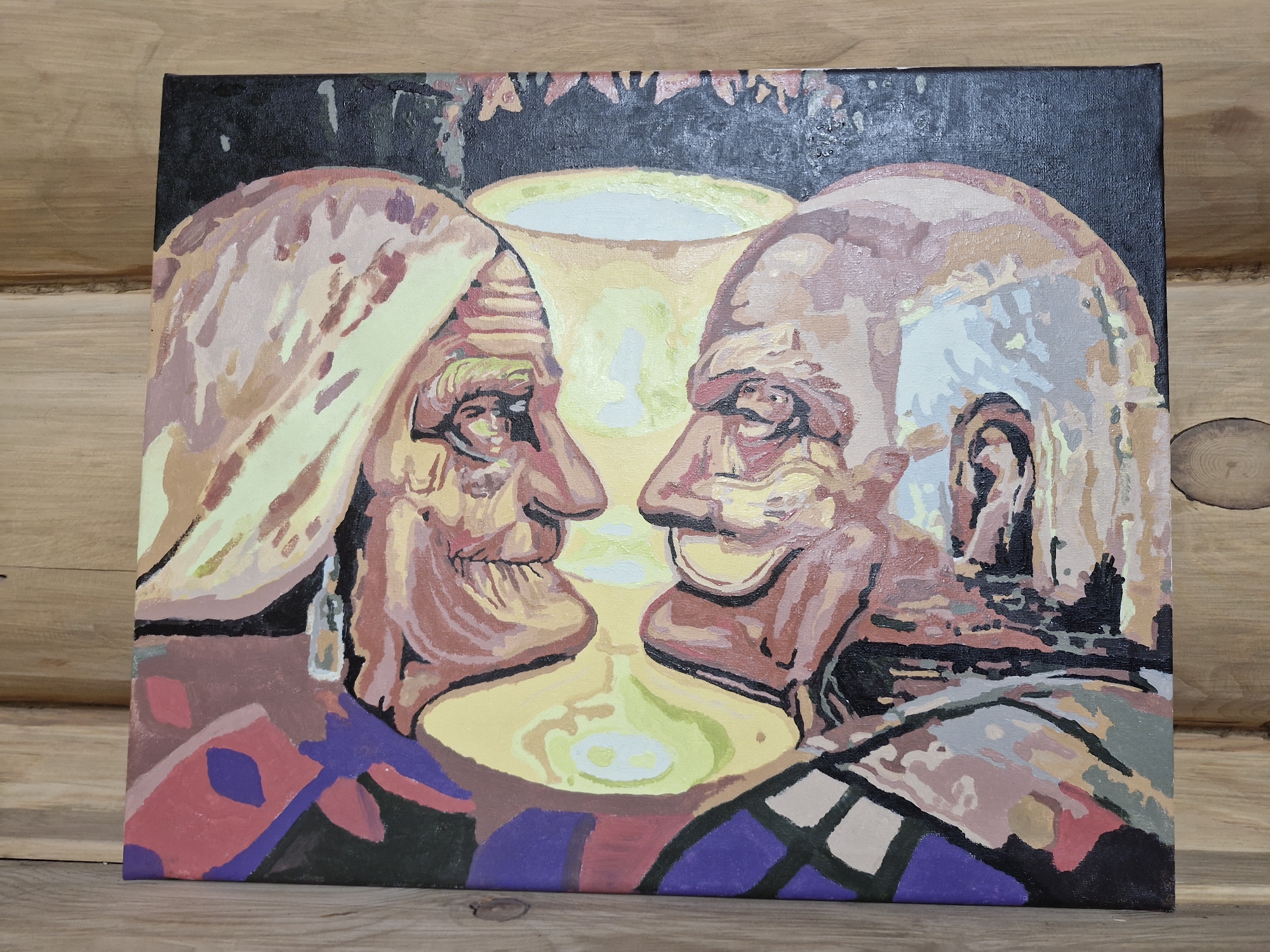 The image depicts a painting on canvas, resting on a wooden surface. The painting is an optical illusion, featuring two large faces in profile facing each other, with their negative space forming a goblet. Within the negative space, two smaller figures are visible, one playing a stringed instrument. The painting uses a palette of earth tones and muted colors, with blocks of purple and red in the foreground. The background is dark and abstract, with some lighter shapes suggestive of foliage or a canopy. The painting is positioned on a wooden surface that appears to be the exterior wall of a structure, likely made of logs, with visible grain and texture. The scene is indoors, with no discernible city or geographical context provided within the image itself. However, the prompt specifies the location as Bulan-Segettu, Kyrgyzstan, implying this is the setting where the painting is displayed, although the image does not offer any visual cues to confirm this.