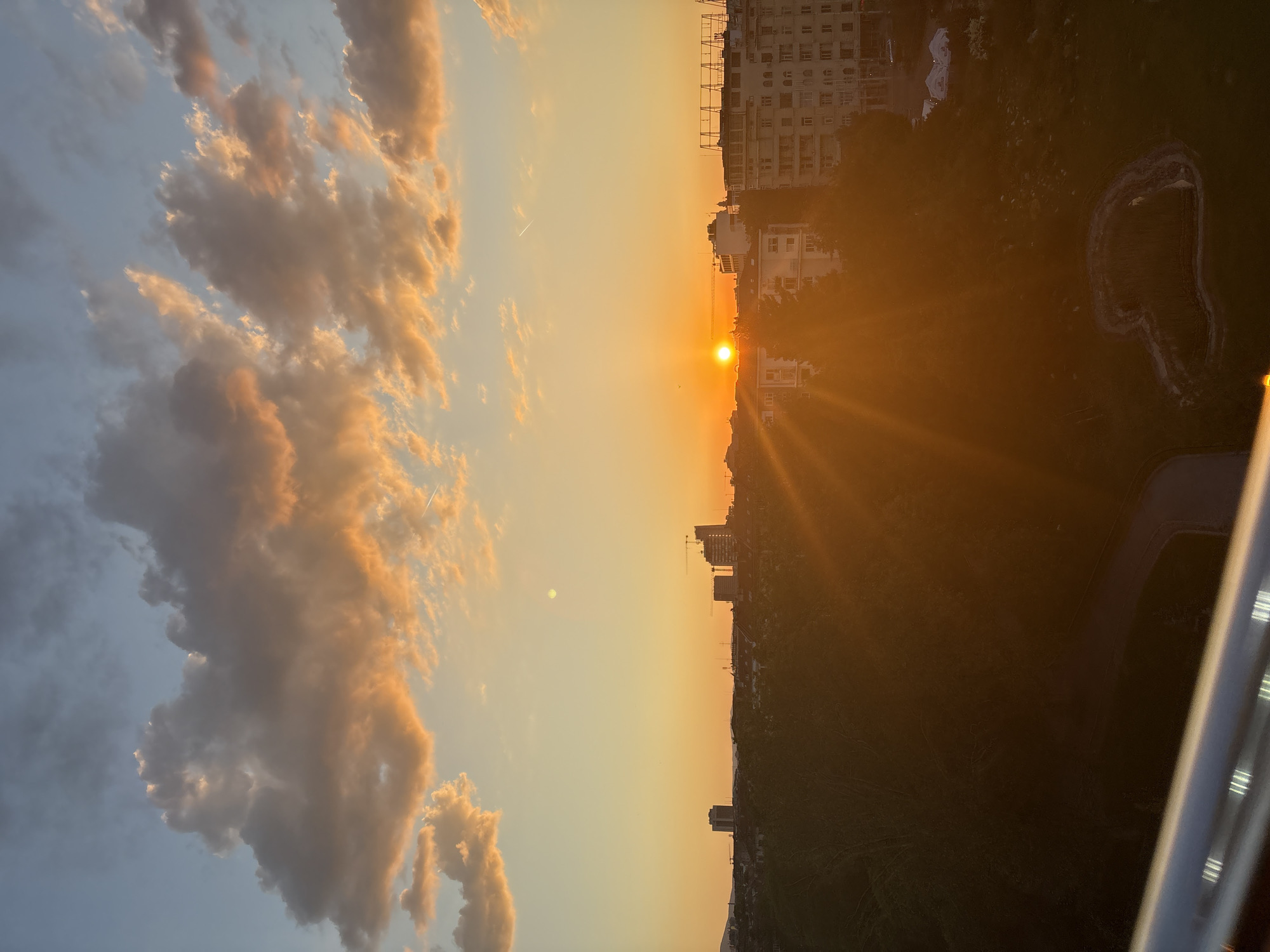 The image captures a sunset over Sofia, Bulgaria. The sun is positioned centrally, emitting a radiant glow that extends across the sky and landscape. Silhouetted against the sun are the city's skyline, including buildings and what appears to be a crane. A green hill slopes away from the urban area. To the left of the sun, clouds are illuminated with orange and yellow hues from the setting sun. The sky transitions from a warm orange near the horizon to a cooler blue at the top left. The lower right corner of the image shows a blurred section of a balcony railing. A small body of water is discernible, reflecting the sunset.