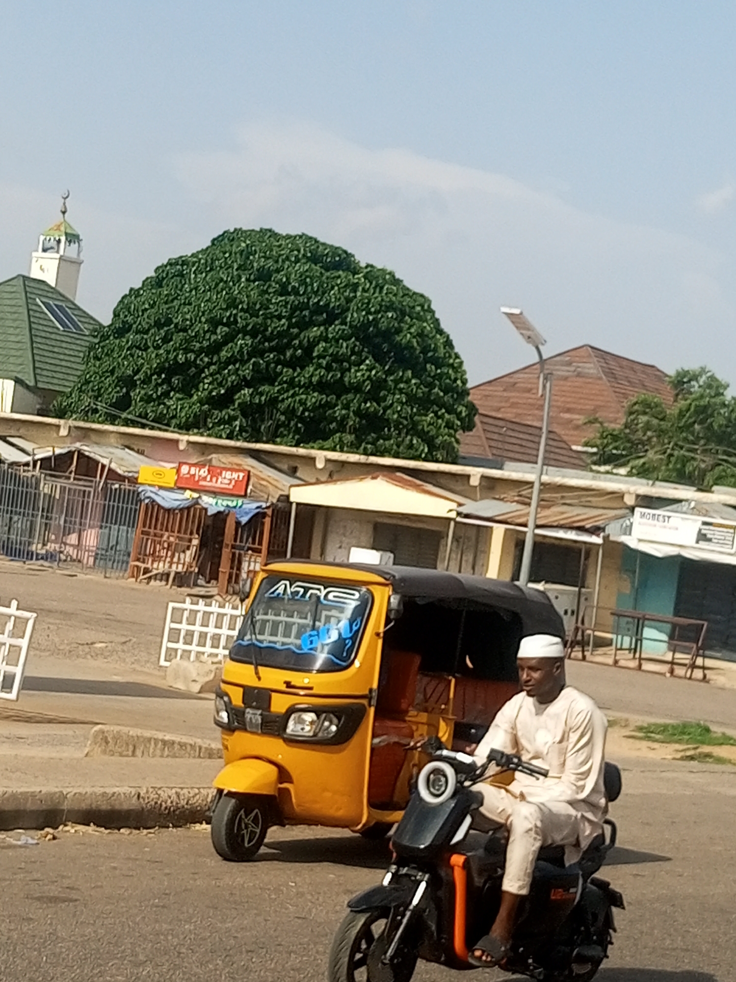 A low-angle, full shot captures a man in a light-colored, traditional robe and white cap riding a black scooter in an urban environment. The scooter is positioned in the foreground, slightly to the right of the frame, with the man looking to his right. Behind him, a yellow auto-rickshaw, also known as a tuk-tuk, is parked, facing left. The auto-rickshaw has the letters "ATG" visible on its windshield.
The background reveals a street scene with a variety of structures. To the left, a distinctive green-roofed building with a solar panel and a minaret topped with a crescent moon suggests a religious building, possibly a mosque. A large, dense green tree with a rounded canopy dominates the middle ground. Further back, there are several stalls or small shops with corrugated metal roofs and open fronts, some with visible signage. One sign reads "MO BEST". A street lamp with a solar panel stands near the shops. The ground appears to be a paved road with some curb and loose gravel. The sky is light blue with some wispy clouds. The overall impression is a snapshot of daily life in a town or city.