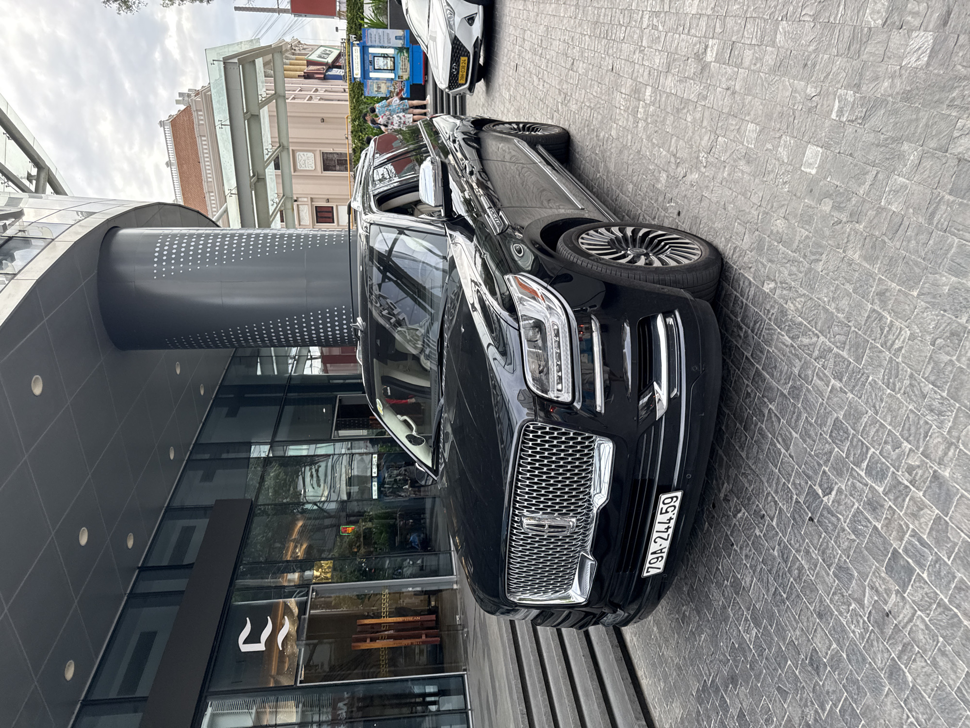 A black Lincoln Navigator SUV is parked on a cobblestone driveway in front of a modern building. The building features a large glass facade and a prominent grey cylindrical pillar with a white dot pattern. The SUV has a chrome grille and a license plate that reads "79A-244.59". To the right of the SUV, a white Hyundai Accent is also parked. In the background, people are visible near a kiosk and in front of a building with traditional architecture. The sky is overcast. The scene appears to be located in Nha Trang, Vietnam, likely at the entrance of a hotel or a commercial establishment.