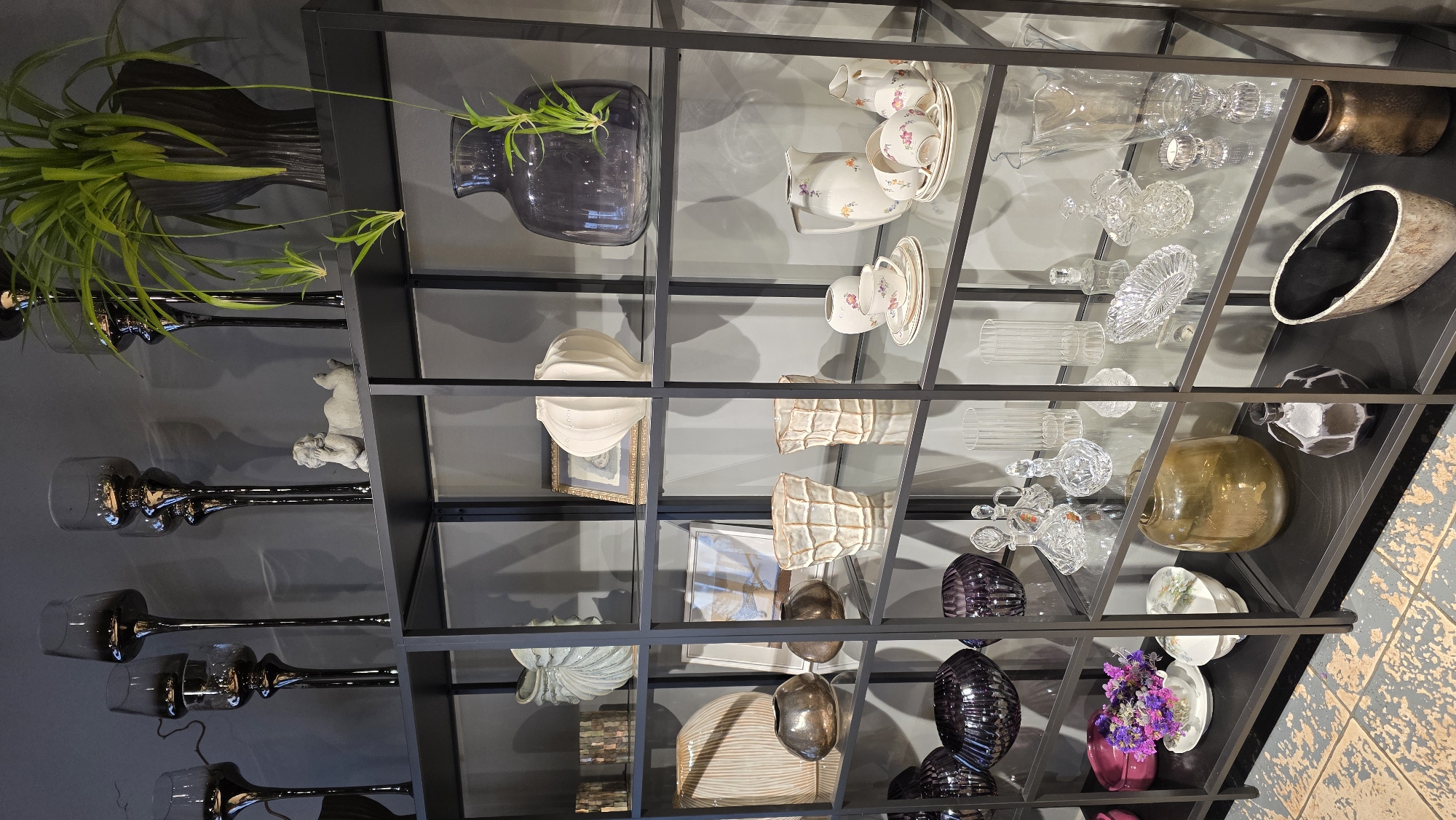 The image shows a display of decorative vases and tableware on a black metal shelf unit. The shelves are made of glass and are filled with various objects, including tall dark grey glass candle holders, a light grey ceramic sculpture of a reclining cherub, a large dark grey vase with a plant, light blue and cream colored vases, framed pictures, and a set of teacups and saucers. The lower shelves contain glass bottles, crystal decanters, a large amber colored vase, a dark patterned vase, and a woven basket. The setting appears to be an indoor retail space or a home decoration display. The flooring in the foreground is a distressed tile pattern. There are no people visible in the image. The scene is lit with artificial light, casting shadows from the objects onto the wall and shelves. There is no information about the location of this scene in Riga, Latvia.