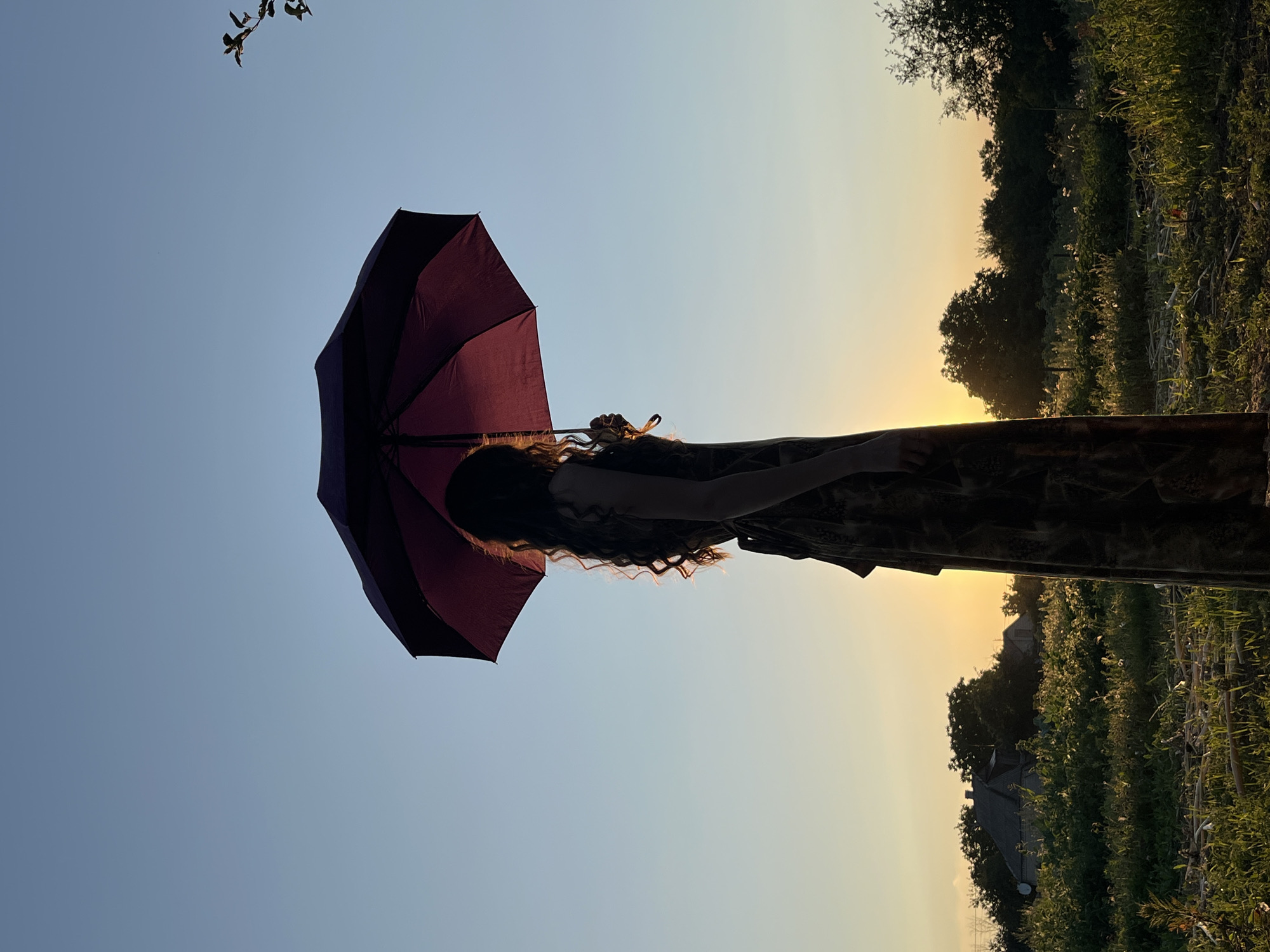 A silhouette of a person stands in a field at sunset, holding an open umbrella above their head. The person is facing right, with long, curly hair illuminated by the setting sun. They are wearing a long, patterned dress. The background shows a rural landscape with trees, houses, and a clear sky transitioning from yellow to blue. The sun is low on the horizon, casting a warm glow.