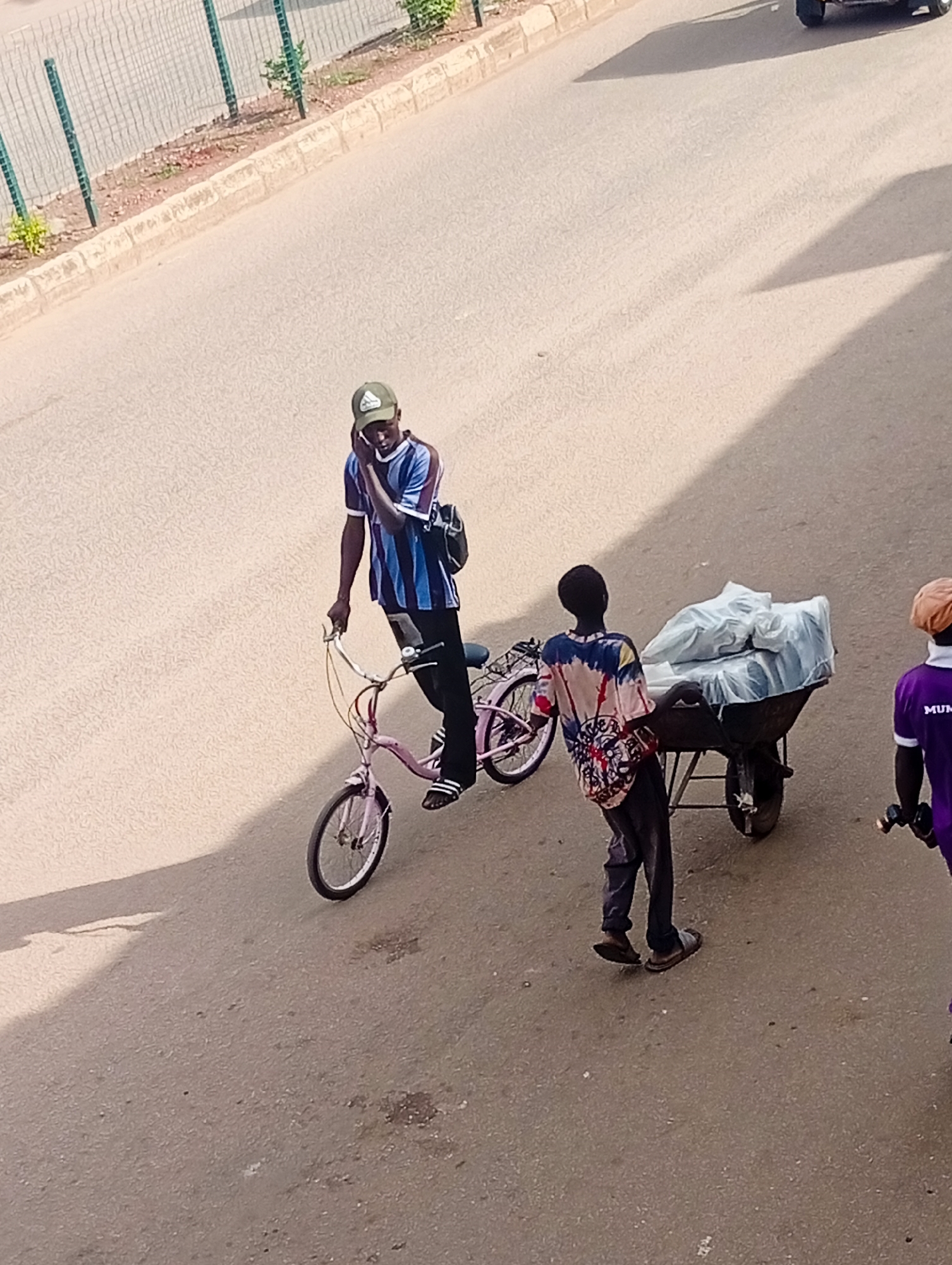 The image captures a street scene in Bukuru, Nigeria, from an elevated perspective. In the foreground, a young man wearing a striped t-shirt, dark pants, and sandals is pushing a wheelbarrow loaded with white-wrapped items. To his left, another young man on a pink bicycle, also in a striped t-shirt, dark pants, and sandals, is holding a phone to his ear. Behind them, a third individual in a purple shirt is partially visible on the right. The environment is an asphalt road with a concrete curb and a metal fence along the side, suggesting an urban or semi-urban setting. The lighting indicates it is daytime, with strong shadows cast by the individuals and their vehicles.