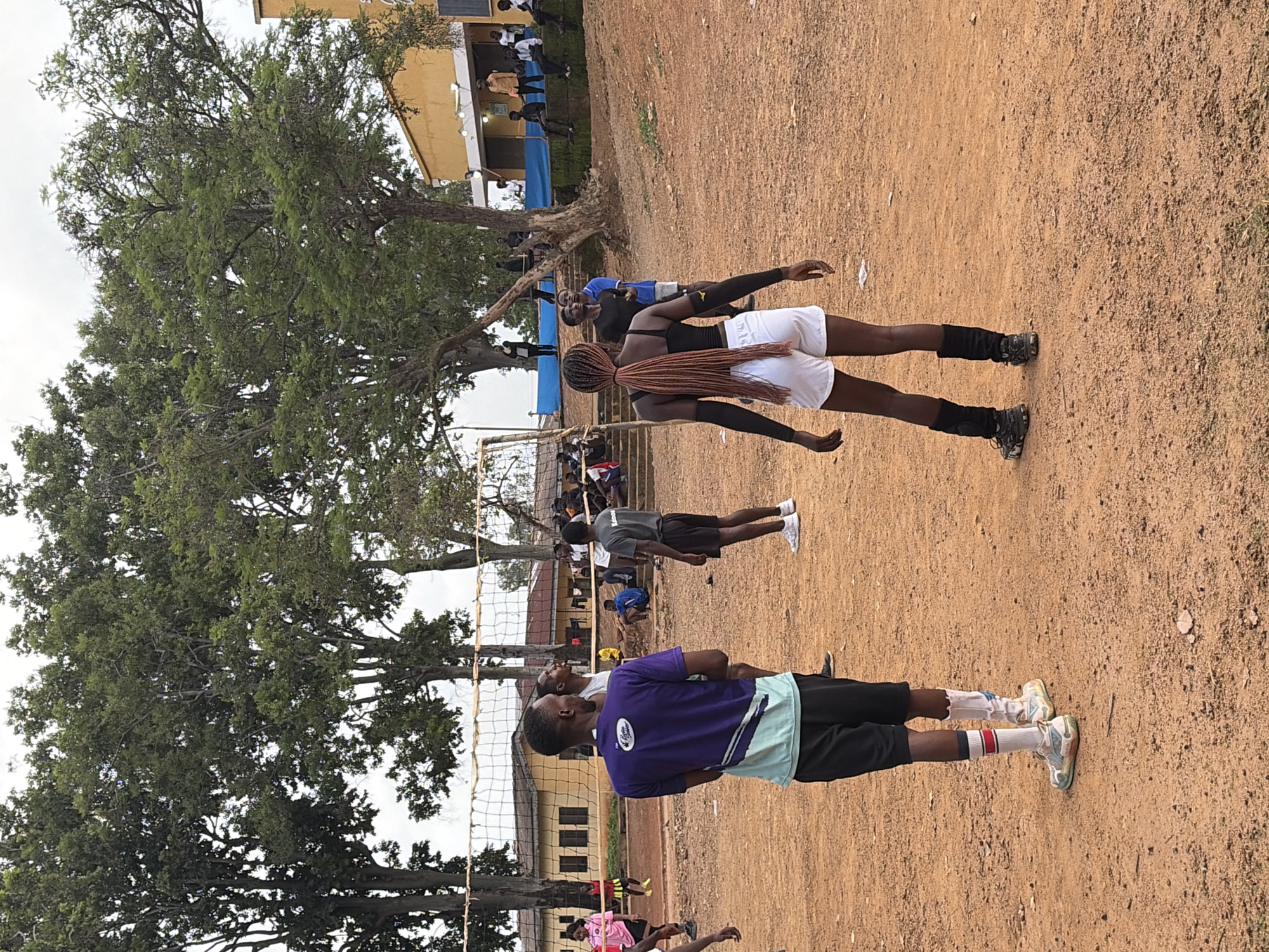 The image depicts a group of young people on an outdoor dirt court, likely a volleyball court, given the presence of a net. The subjects are predominantly young adults, dressed in athletic wear.
In the foreground, a person wearing a purple t-shirt with a light blue and white trim and black shorts is facing away from the camera. They have white socks with red stripes and colorful athletic shoes. Behind them, another individual in a black tank top and white shorts, with their hair in long braids, is also facing away. Further back, several other individuals are visible, some in athletic attire, others in more casual clothing, gathered around the court area.
The scene appears to be an outdoor practice or game. The ground is a reddish-brown dirt, typical of many sports fields in Nigeria. Lush green trees with dense foliage frame the scene, suggesting a tropical or sub-tropical environment. In the background, there's a building with a yellow facade and a shaded overhang, possibly a school or community center. The sky is overcast, indicating a cloudy day.
The activity appears to be a sports-related gathering, likely volleyball given the court setup. The focus on the individuals in the foreground suggests a moment of preparation or observation. There is no strong indication of specific emotions, but the overall atmosphere seems relaxed and focused on the activity.
The text "Rafin Par, Nigeria" provides the geographical context for this scene. No other visible text is discernible in the image. The time of day appears to be daytime, based on the ambient light, but the overcast sky makes it difficult to pinpoint precisely.